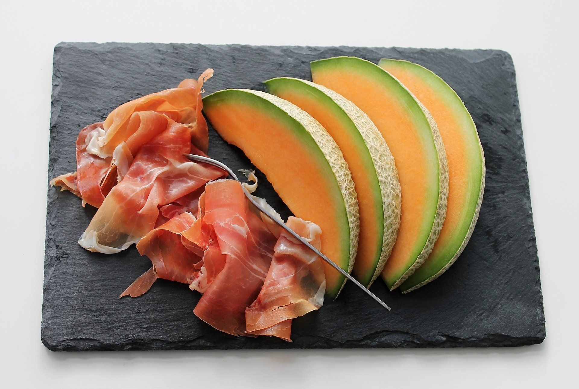 Sliced cantaloupe and prosciutto arranged on a black slate serving board