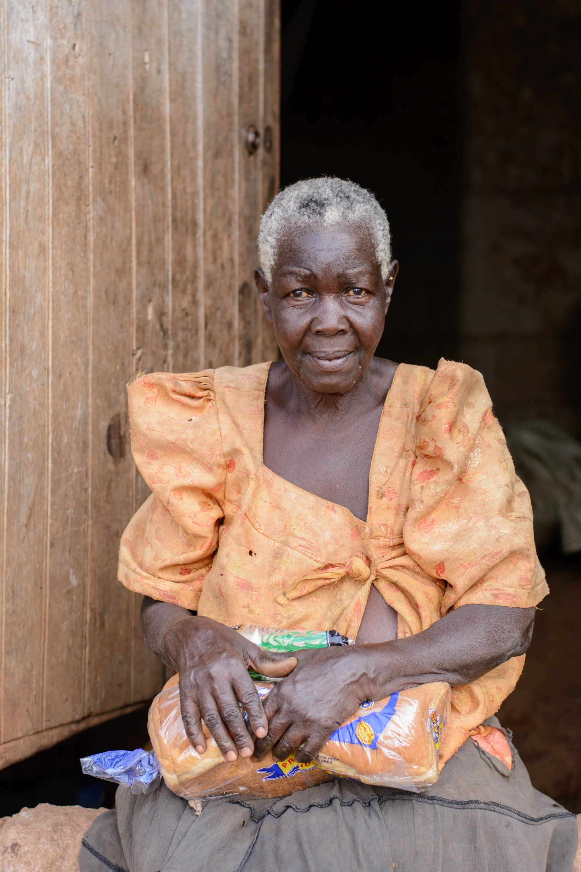 An elderly woman is sitting in front of a wooden door holding a loaf of bread.
