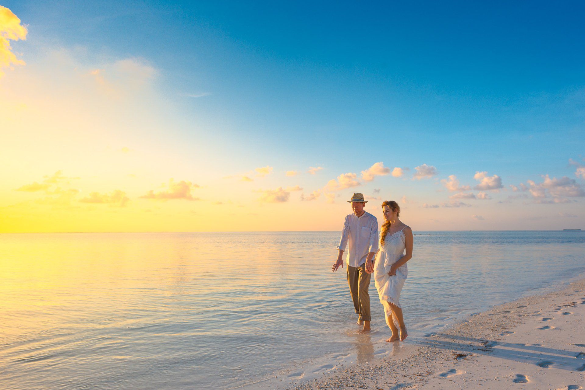 Couple walking barefoot on a beach at sunset, water reflecting sky's colors. Honeymoon holiday