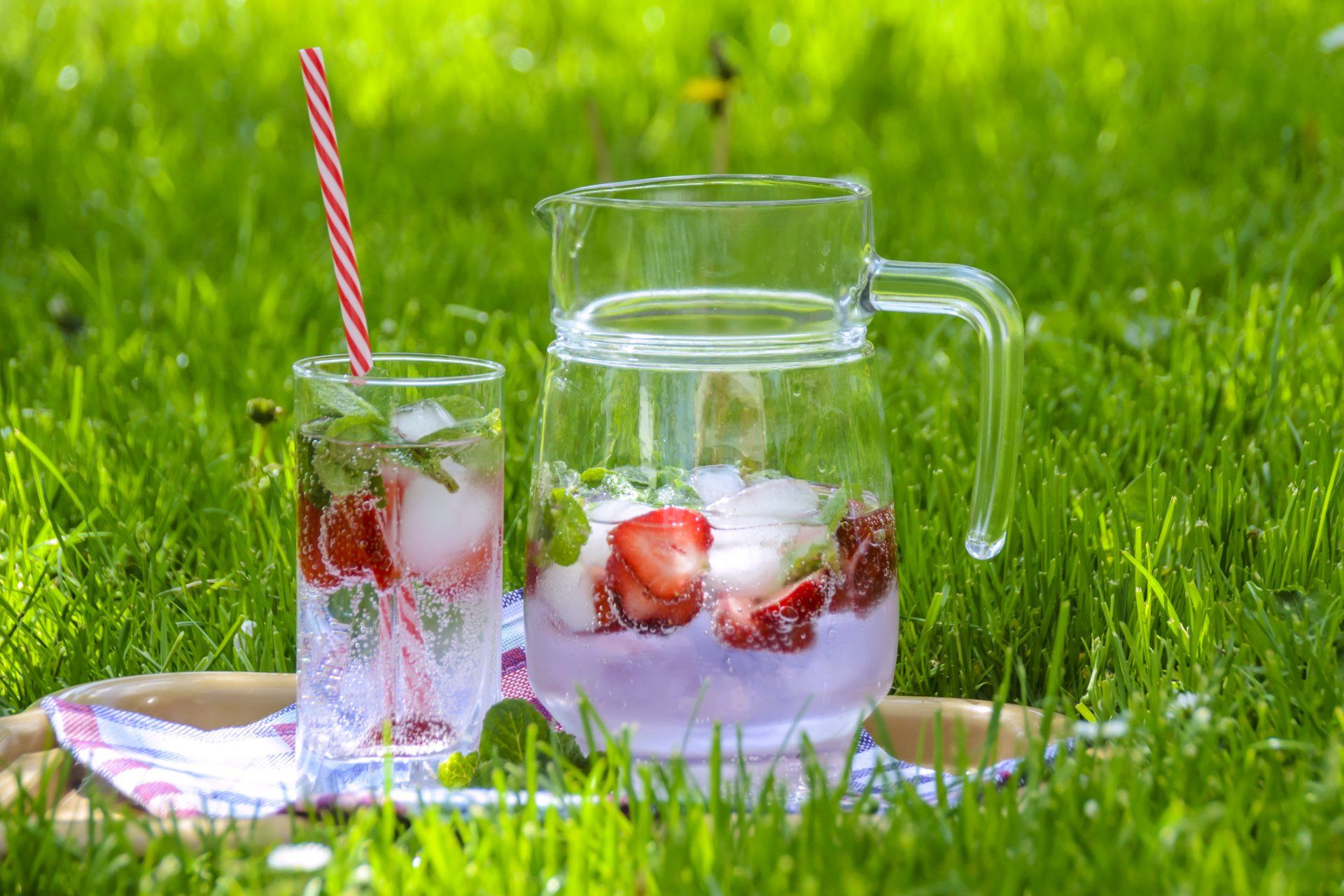 A glass jug of drink with ice and strawberries in with a glass next to it outside on the grass