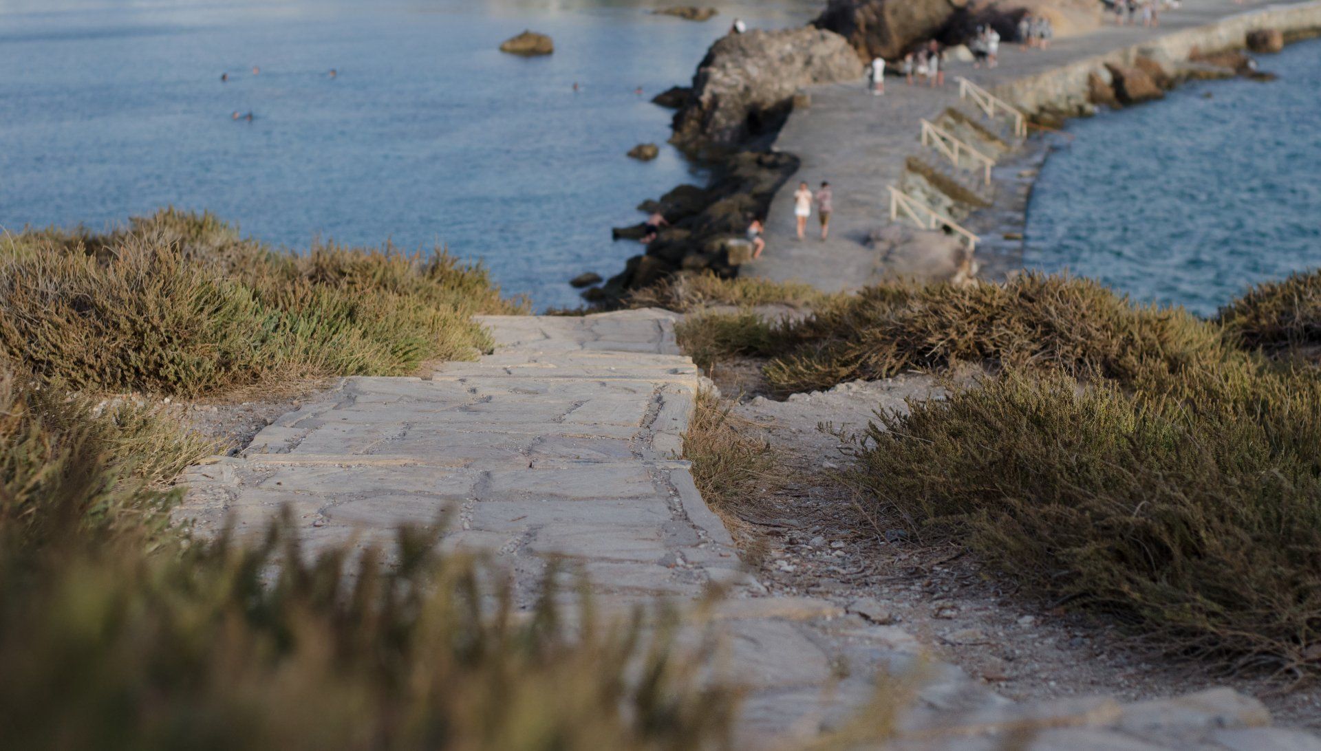 Stone path leading down to a pier with people, surrounded by dry brush, sea in the background.