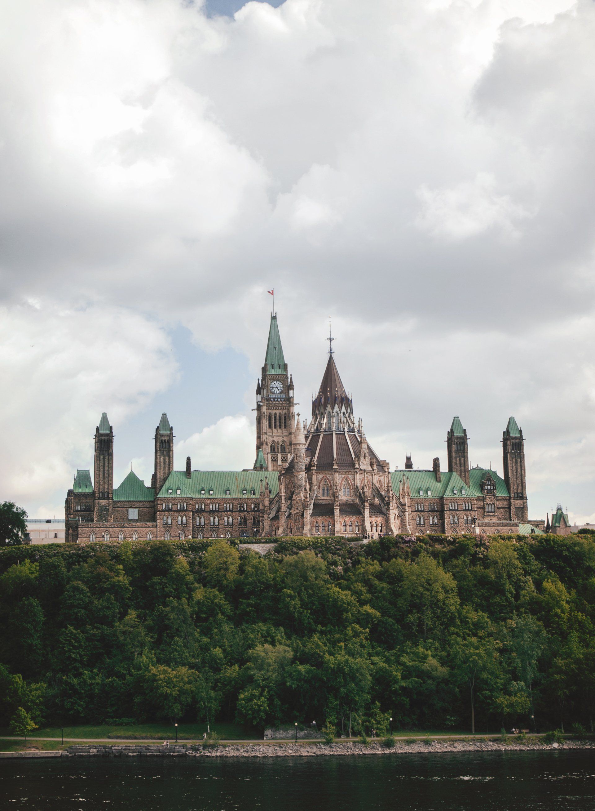 Parliament building with green copper roofs above a tree-lined riverbank under cloudy skies