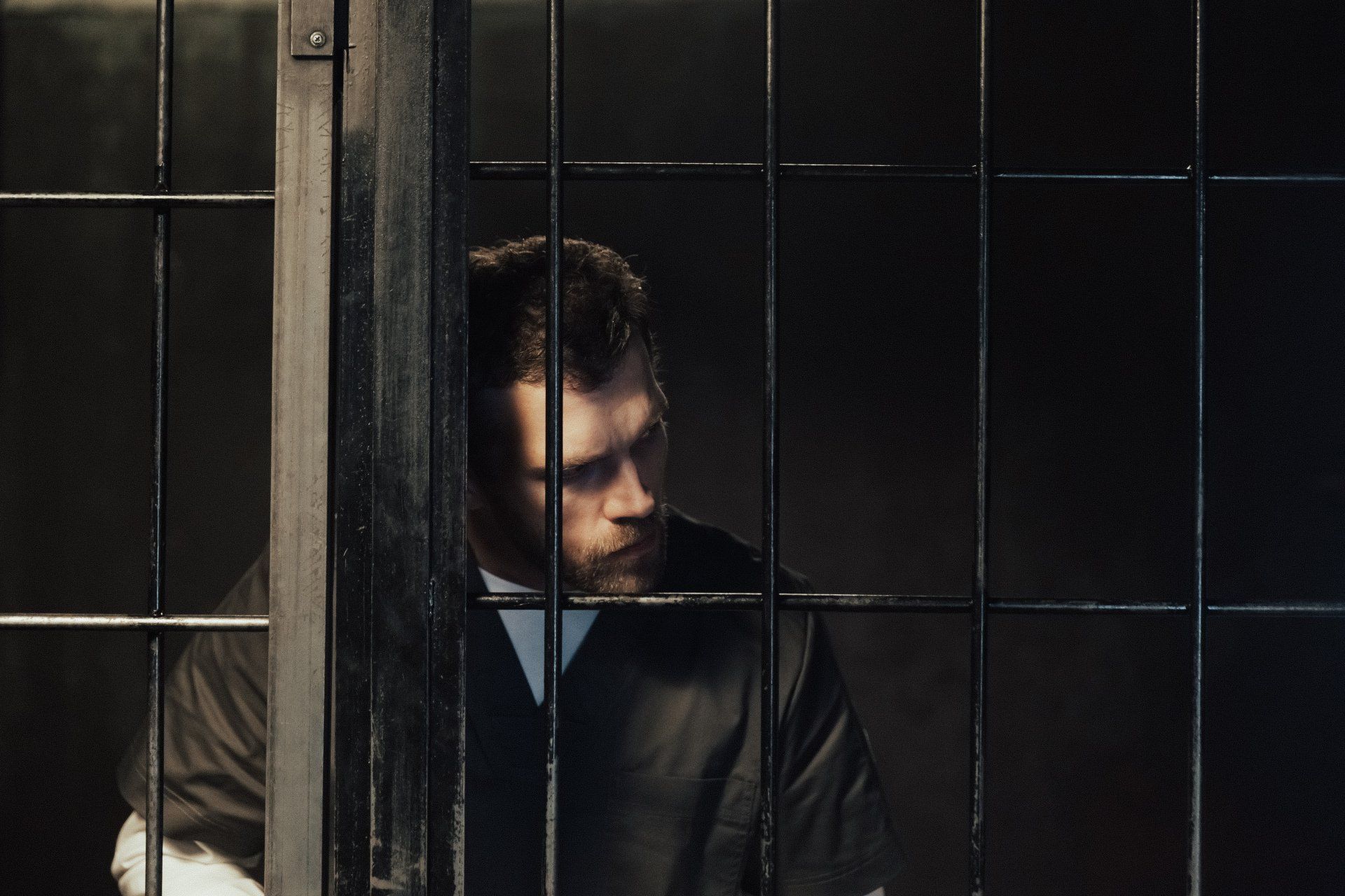 A man in a dark uniform looks toward the side while sitting behind black metal prison bars.