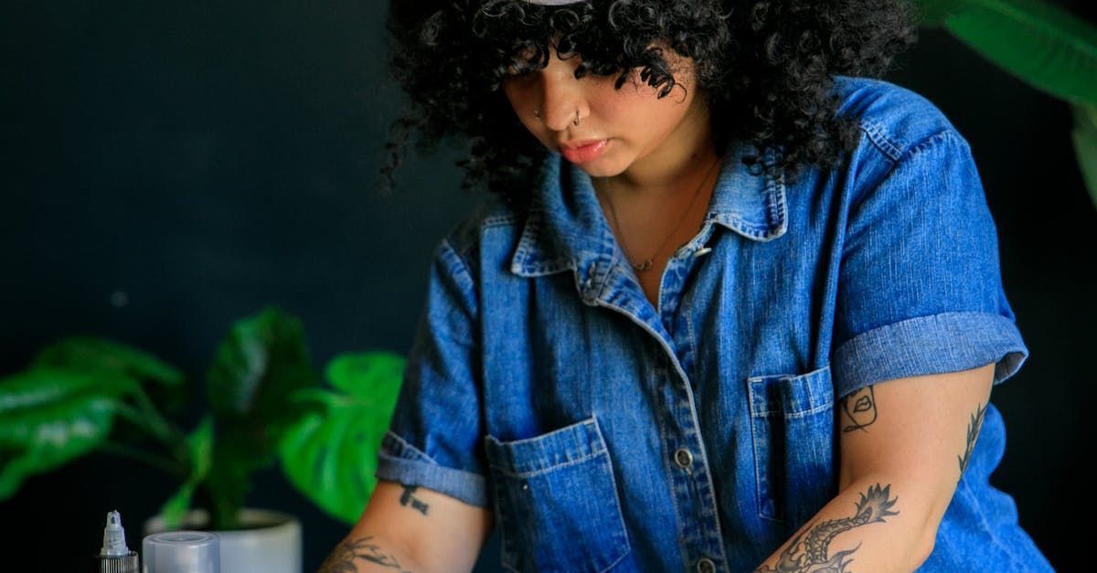A woman with curly hair and tattoos is sitting in front of a potted plant.