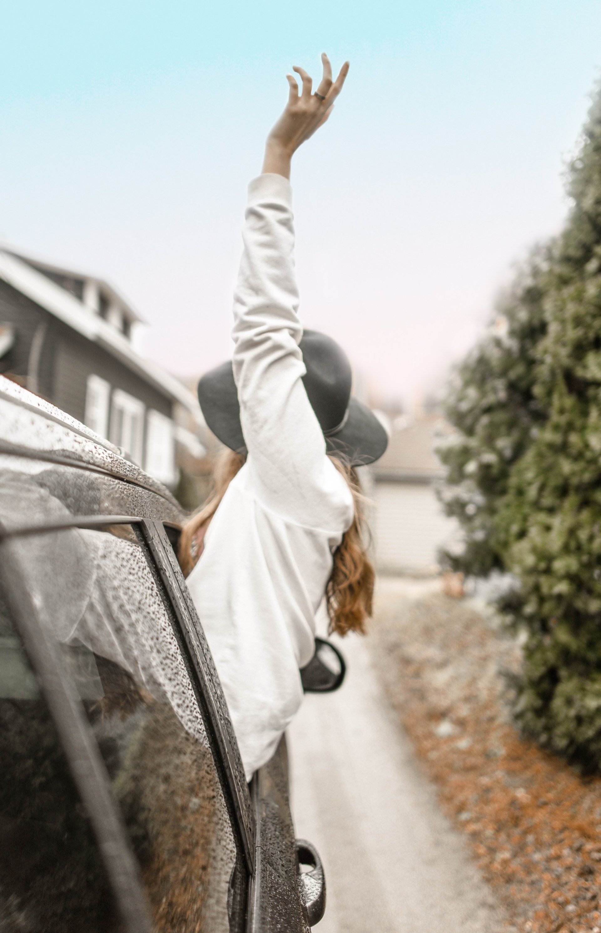 Woman with arm raised out of car window, wearing a hat and white sweater.