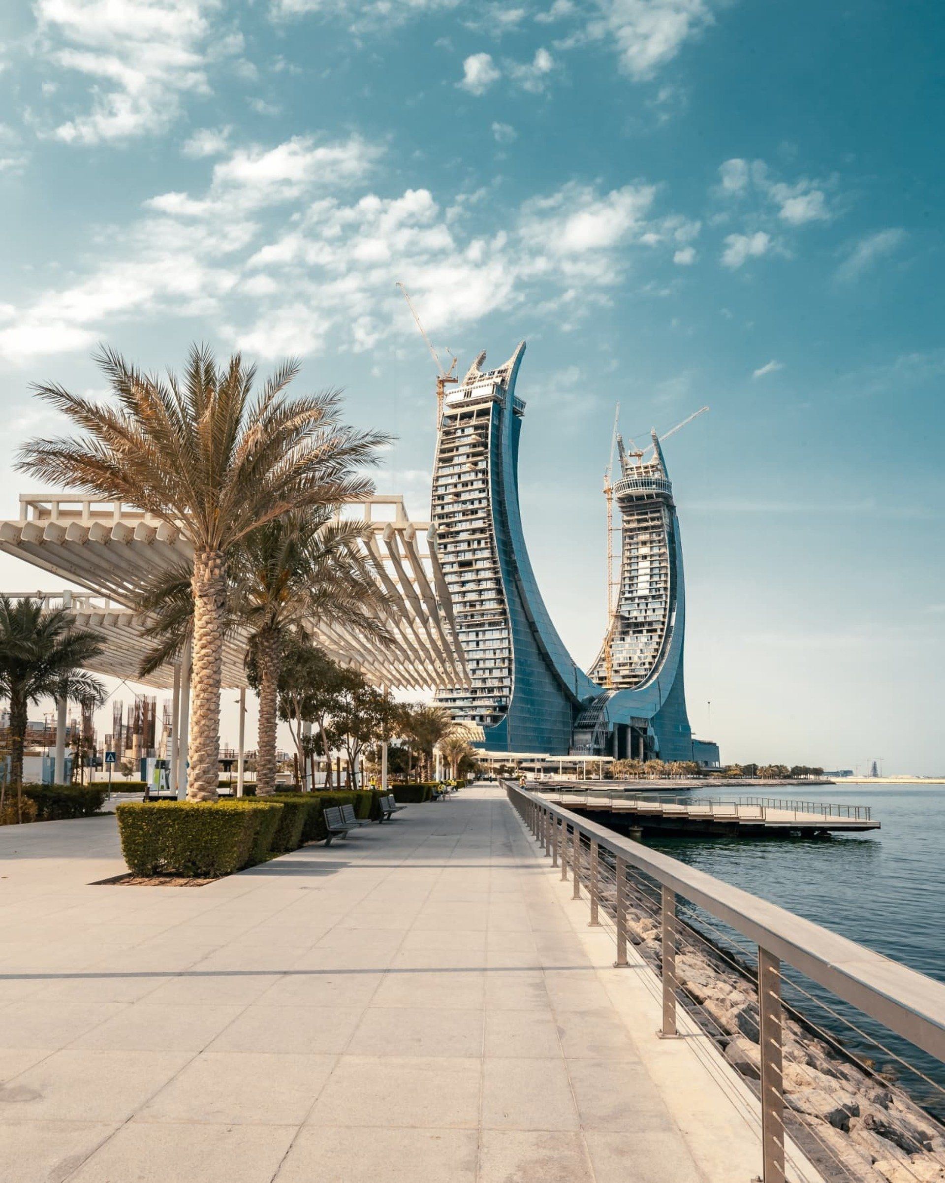 A walkway along the water with palm trees and a large building in the background.