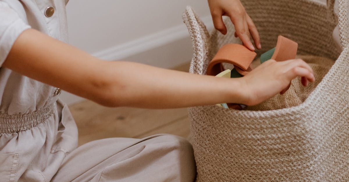 Een persoon in een neutrale outfit reikt in een gevlochten, beige mand met textuur, waarin kleurrijke, gebogen stapelblokken liggen.