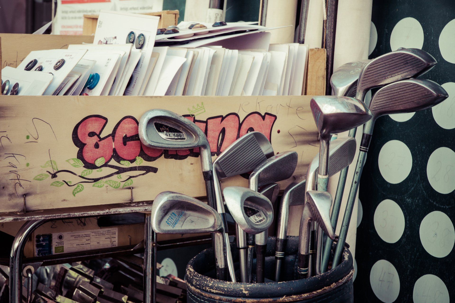 Golf clubs and other equipment in a cluttered store display with graffiti and polka-dot fabric backdrop