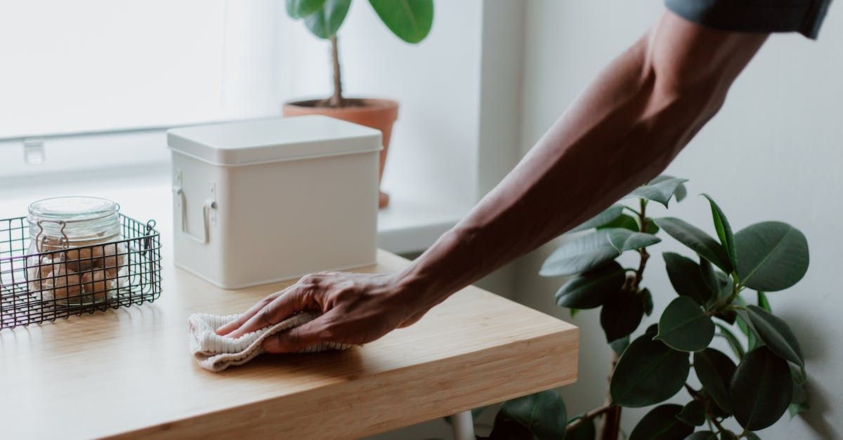 A person wipes down a light wooden desk with a cloth, with a white container and a small plant in the background.