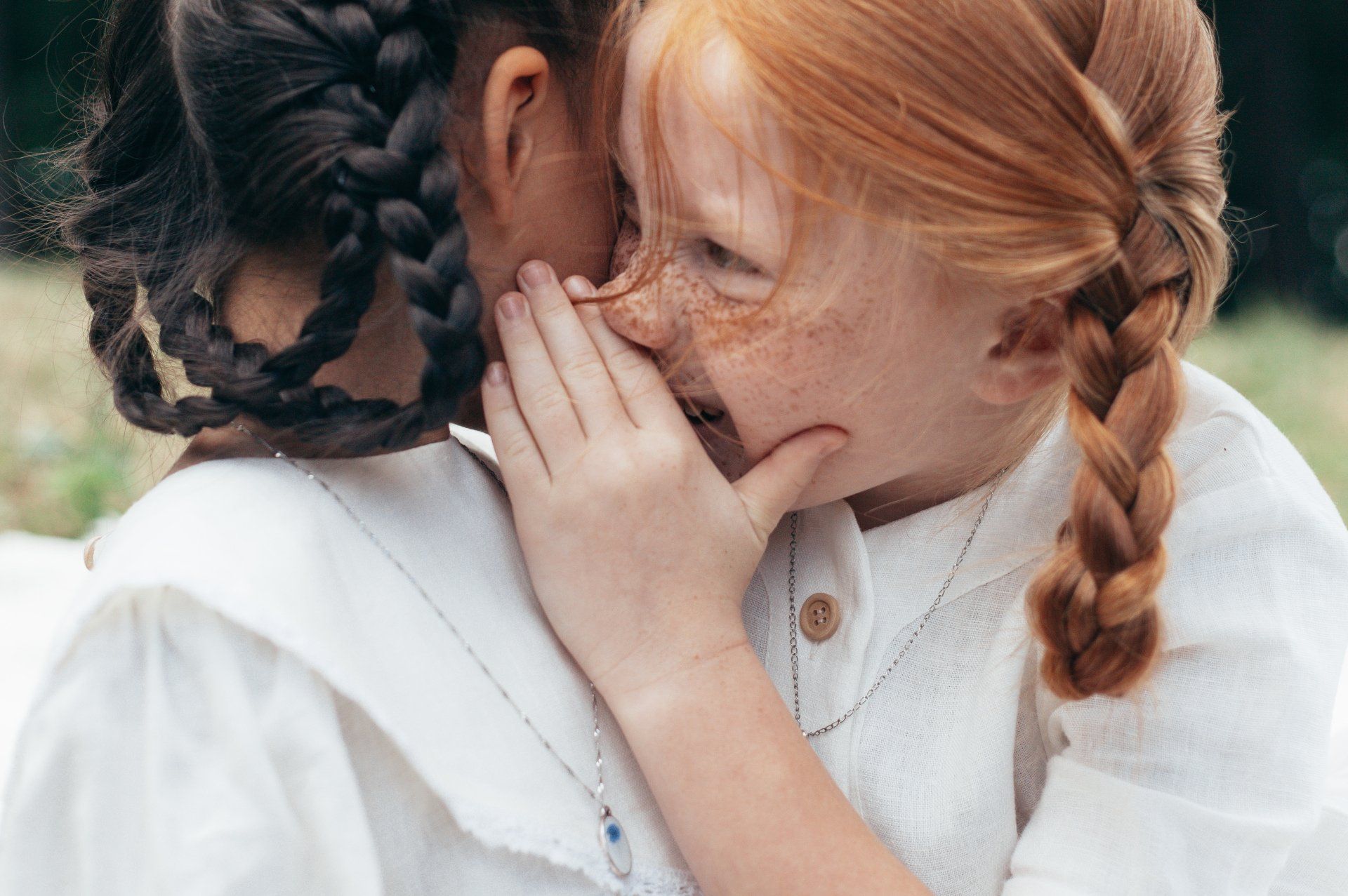 Two children in white shirts whispering close together outdoors