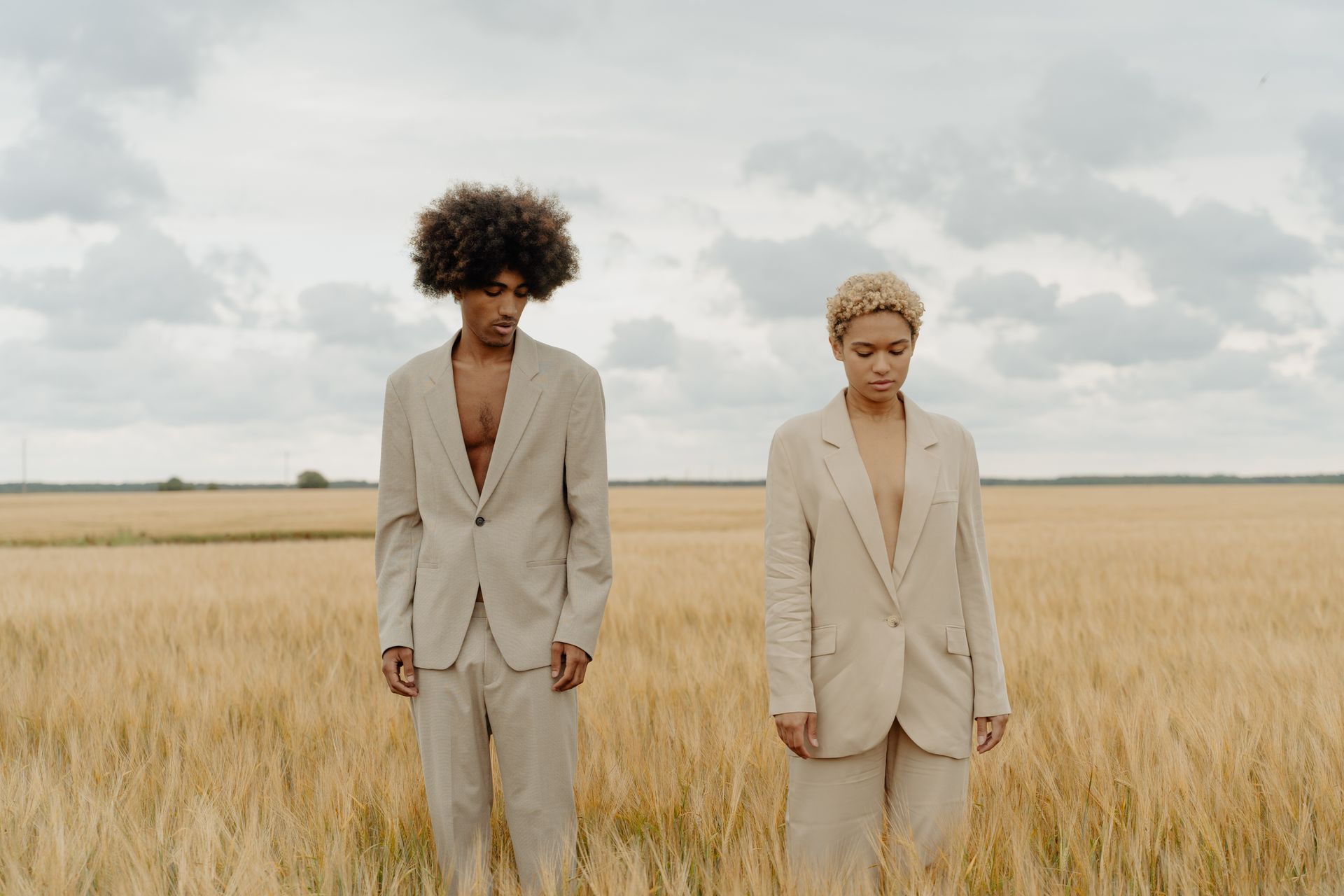Two people in beige suits stand in a wheat field, looking down. Cloudy sky.