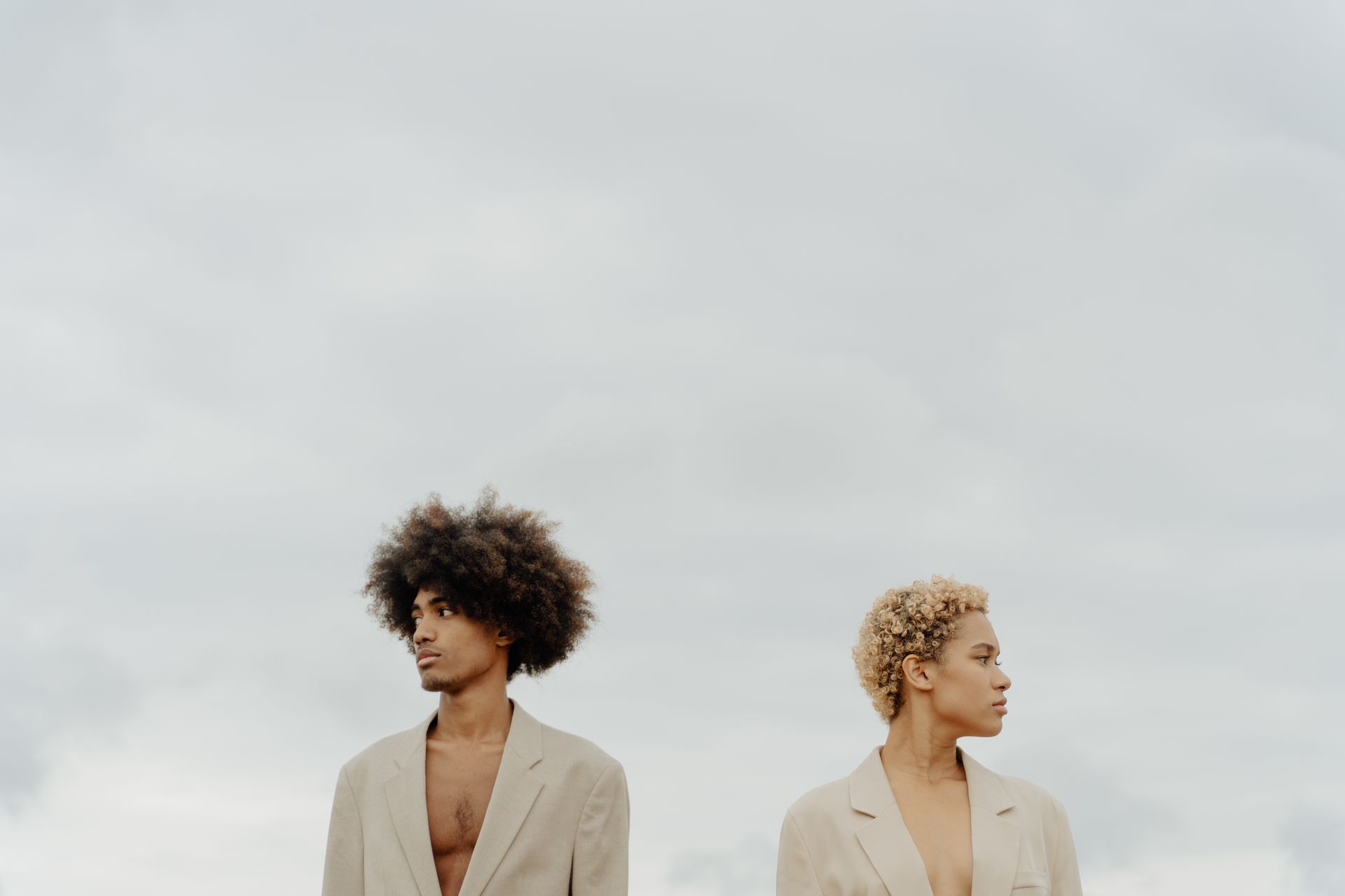 Two people wearing tan blazers, looking away. Cloudy sky backdrop.