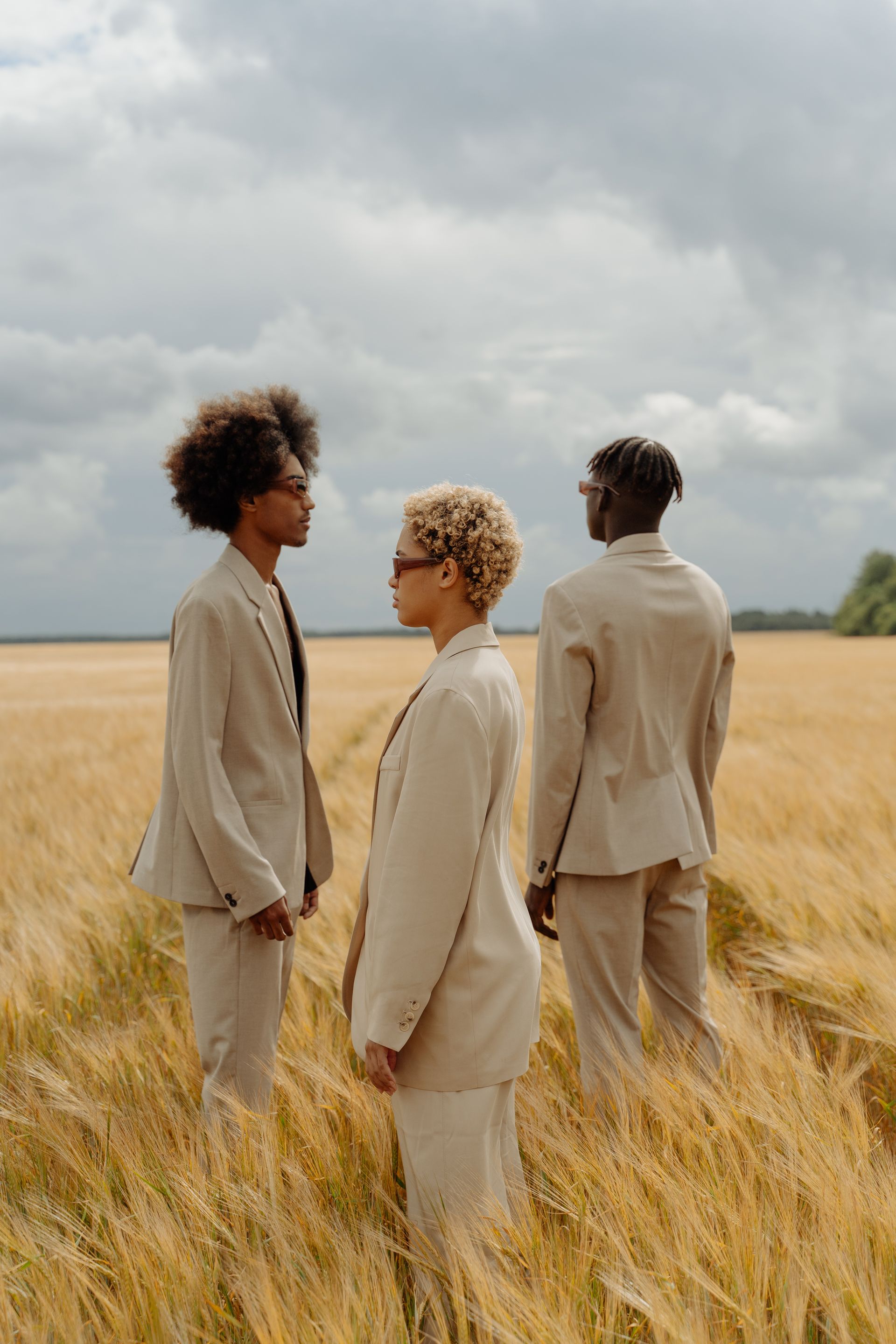 Three people in tan suits stand in a golden wheat field under a cloudy sky.