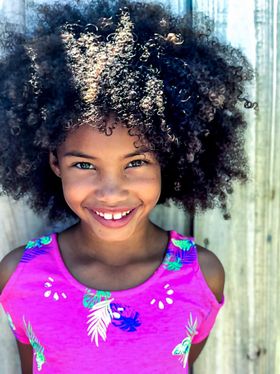 A young girl with curly hair is wearing a pink shirt and smiling.