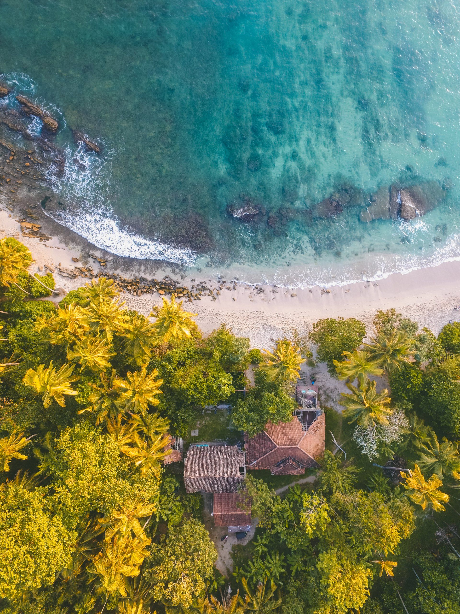 Aerial view of a tropical beach with turquoise water, white sand, and palm trees surrounding small huts.