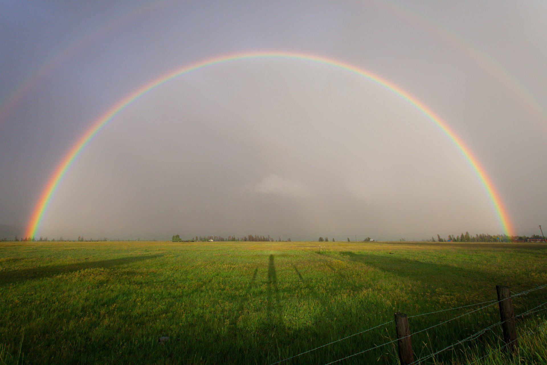 Rainbow arches over a green field and a fence. The rainbow's colors are vibrant.