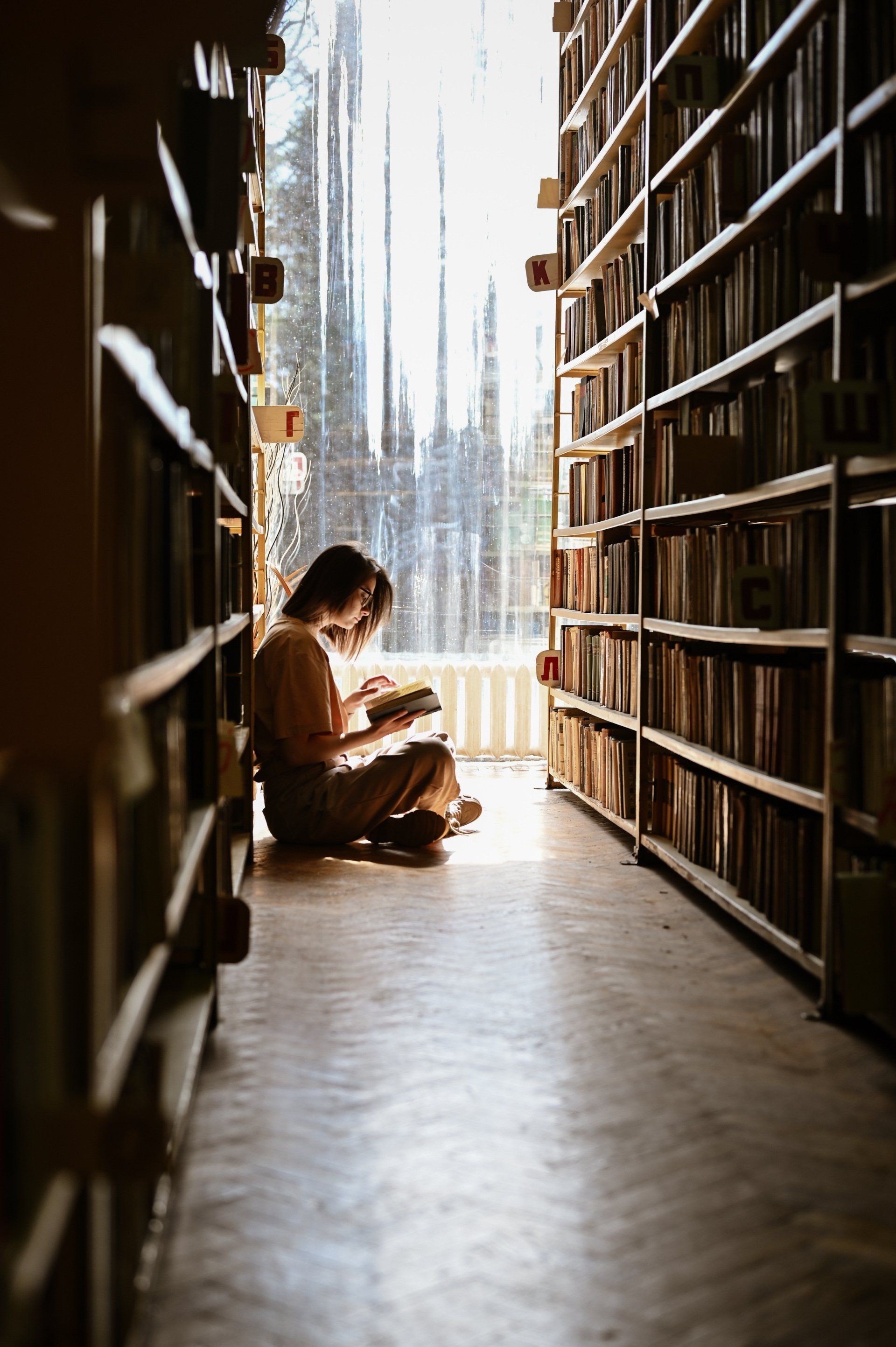 A woman is sitting on the floor in a library reading a book.