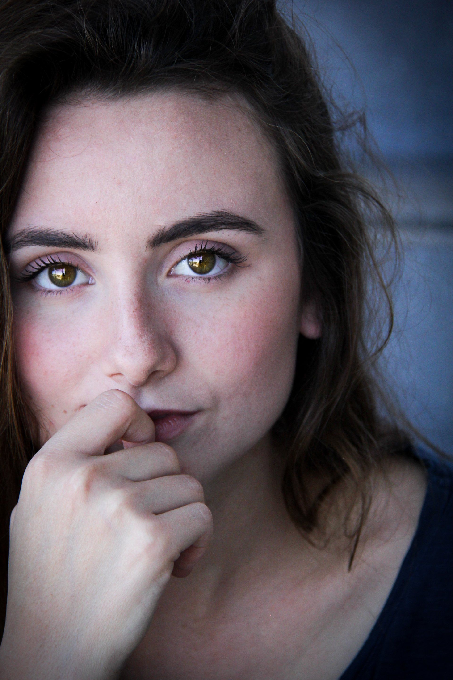 Close-up portrait of a woman with dark hair and green eyes, resting her hand near her mouth.