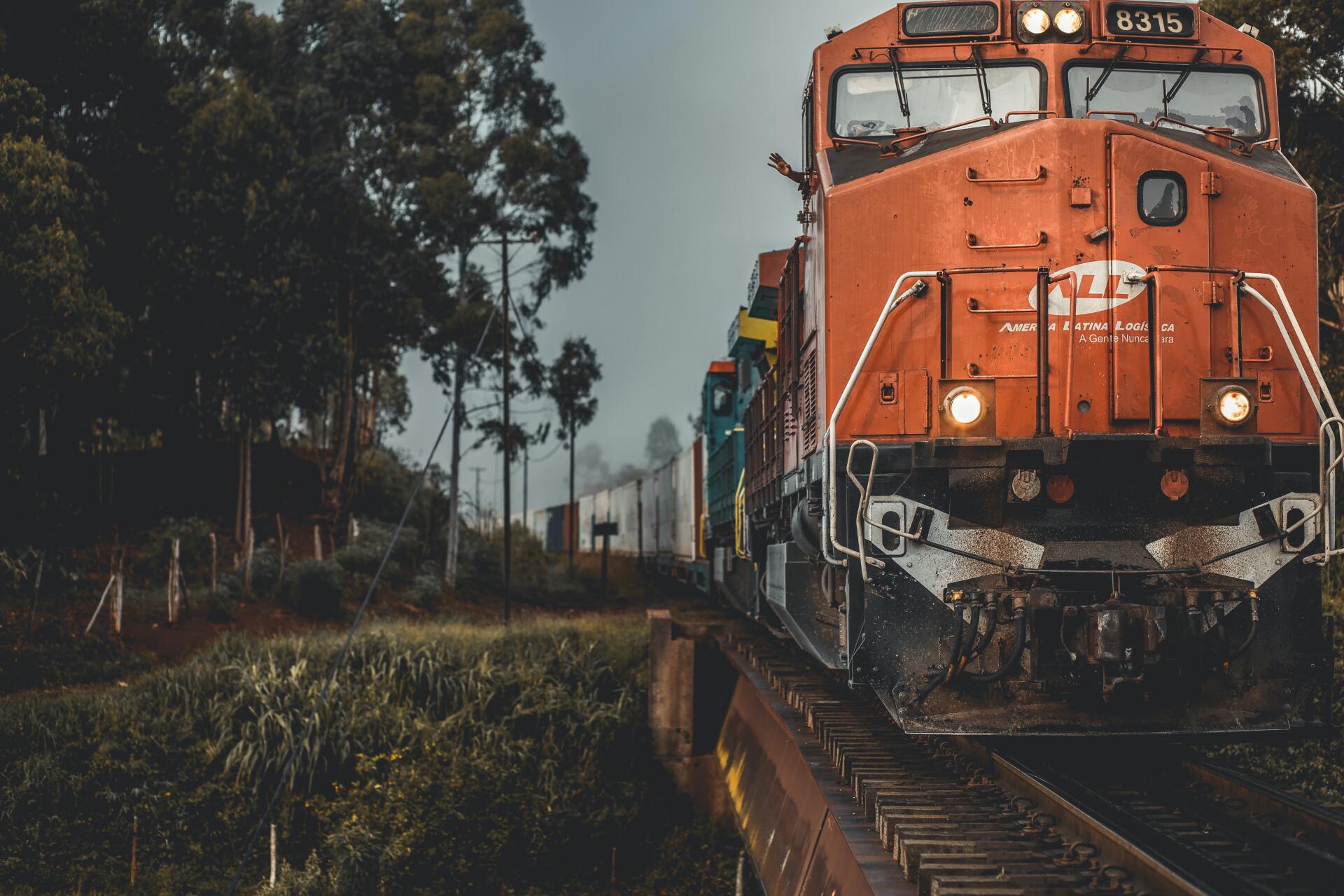 Orange train engine traveling on tracks in a wooded area on an overcast day.