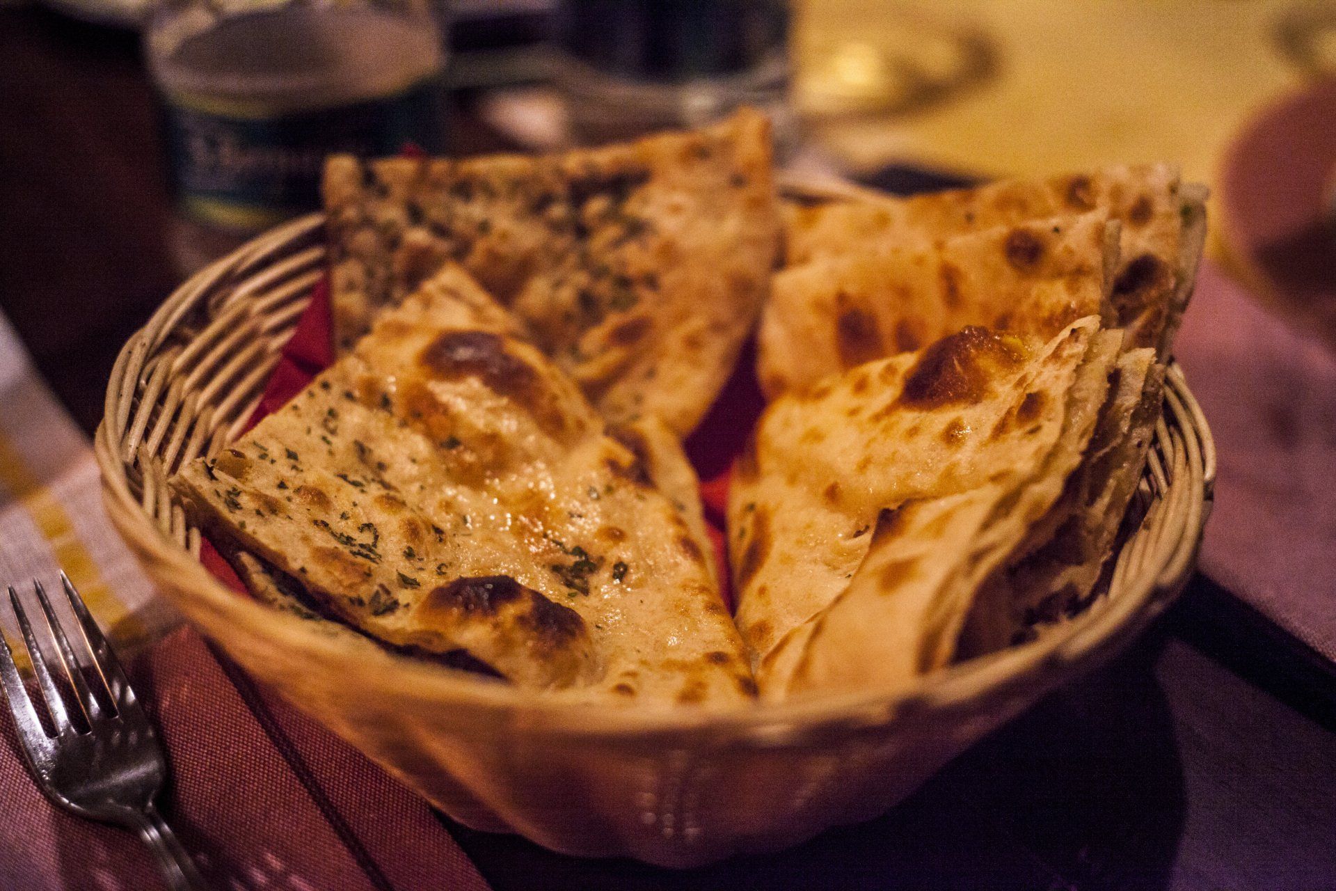 Basket of naan bread, some with herbs, on a table setting with a fork and cloth napkin.