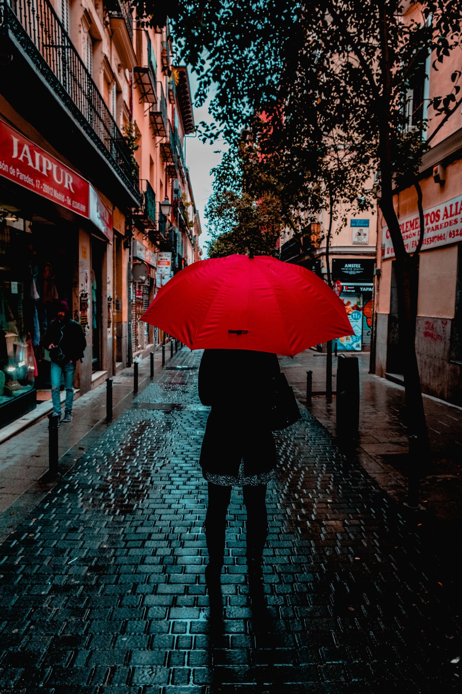 Person with red umbrella walking on wet cobblestone street, flanked by shops and buildings.
