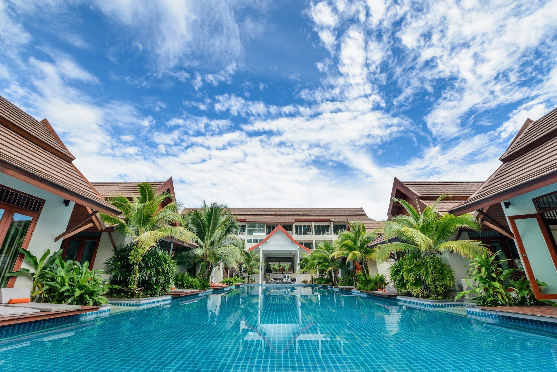 Swimming pool at a resort with surrounding buildings, palm trees, and a cloudy blue sky.