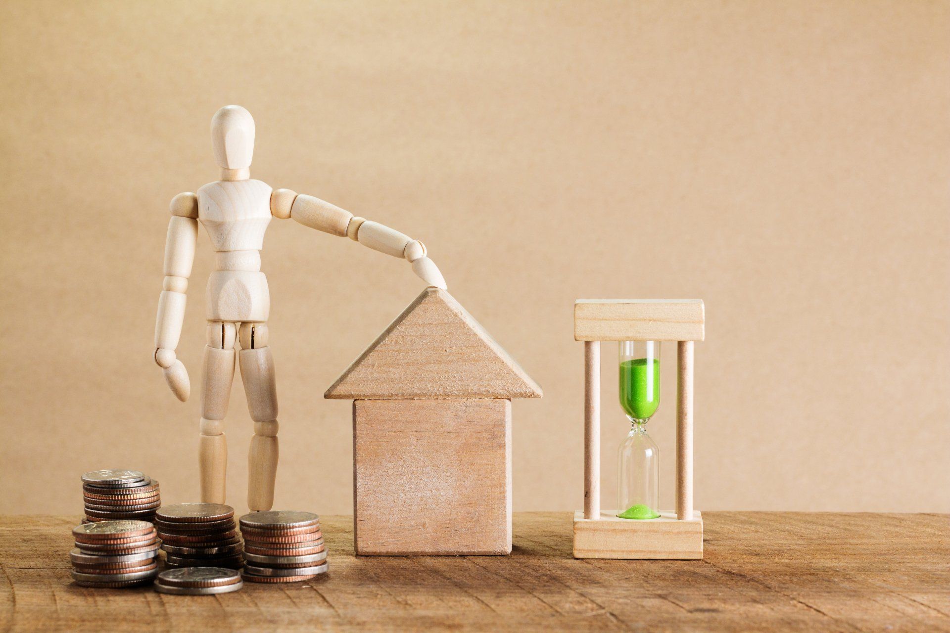 Wooden figure with arm on top of a small wooden house. Wooden hourglass. Small pile of coins in front of wooden figure.