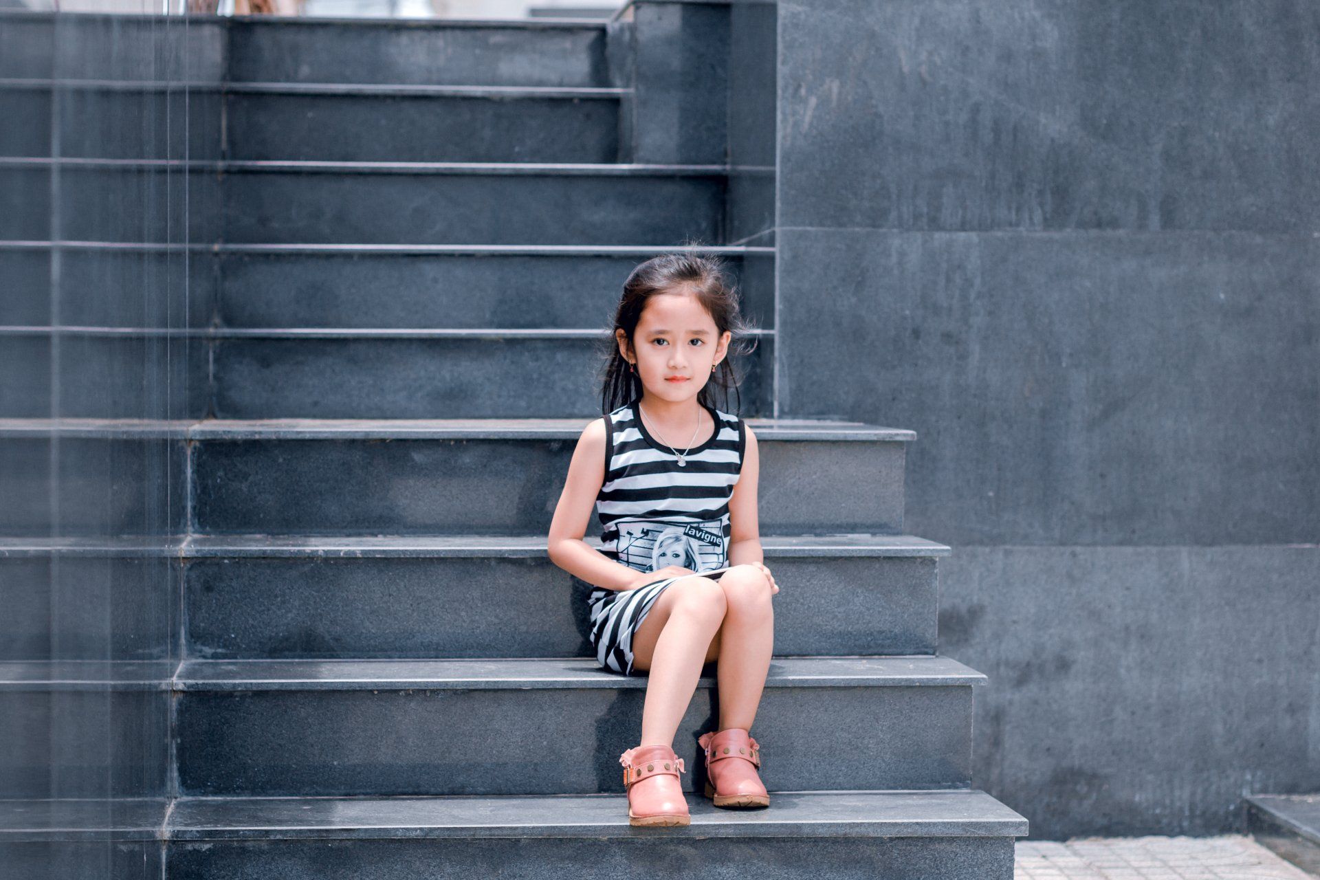 Young child in striped outfit sits on gray steps, looking at the camera.
