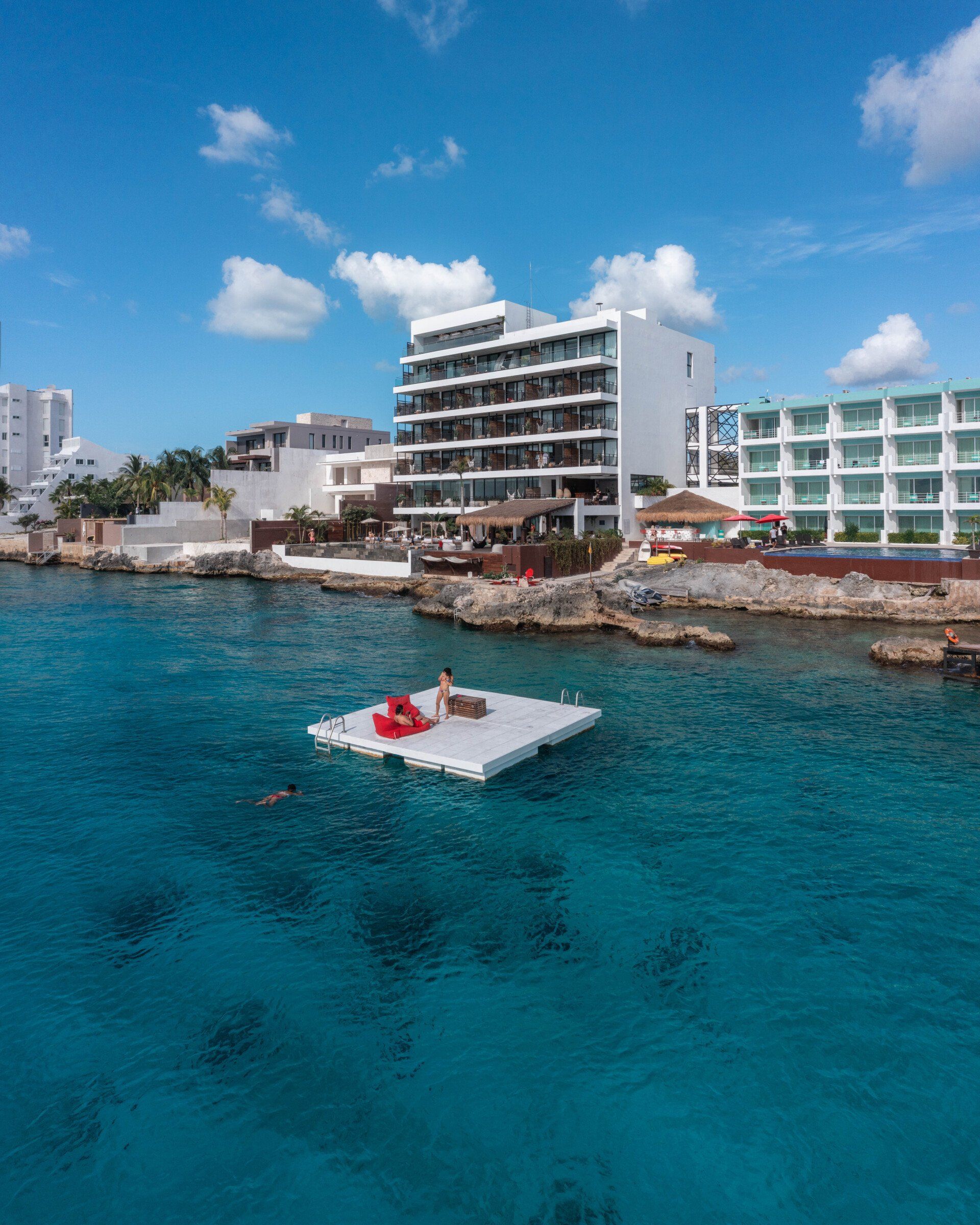 Scroll naar beneden voor het verhaal Drijvende steiger met rode stoelen in turquoise water, gebouwen op de achtergrond onder een blauwe hemel.