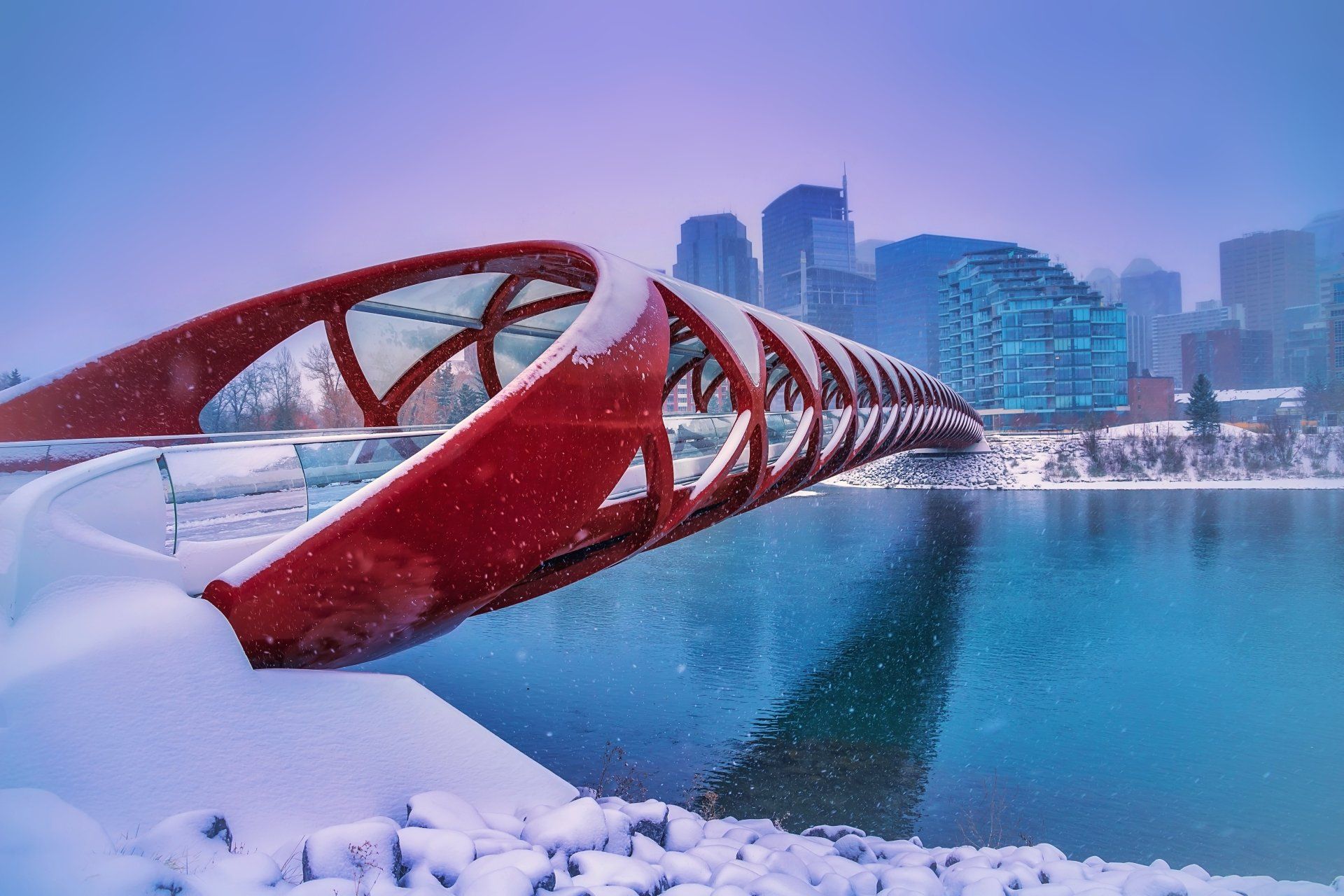 Red Peace Bridge spanning a turquoise river with snow, buildings in the background.