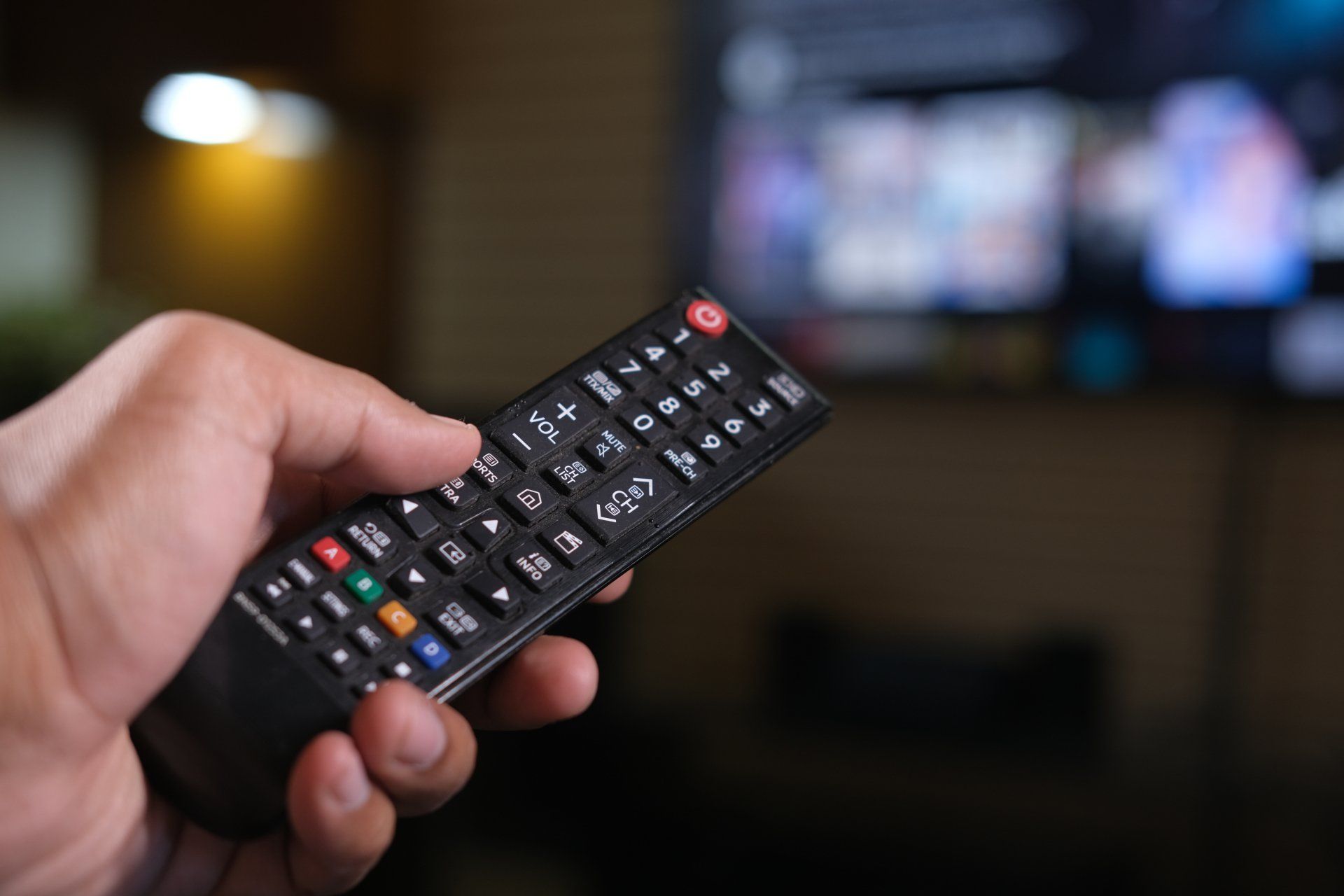 Hand holding a TV remote, pointing it at a blurred television in a dimly lit room.