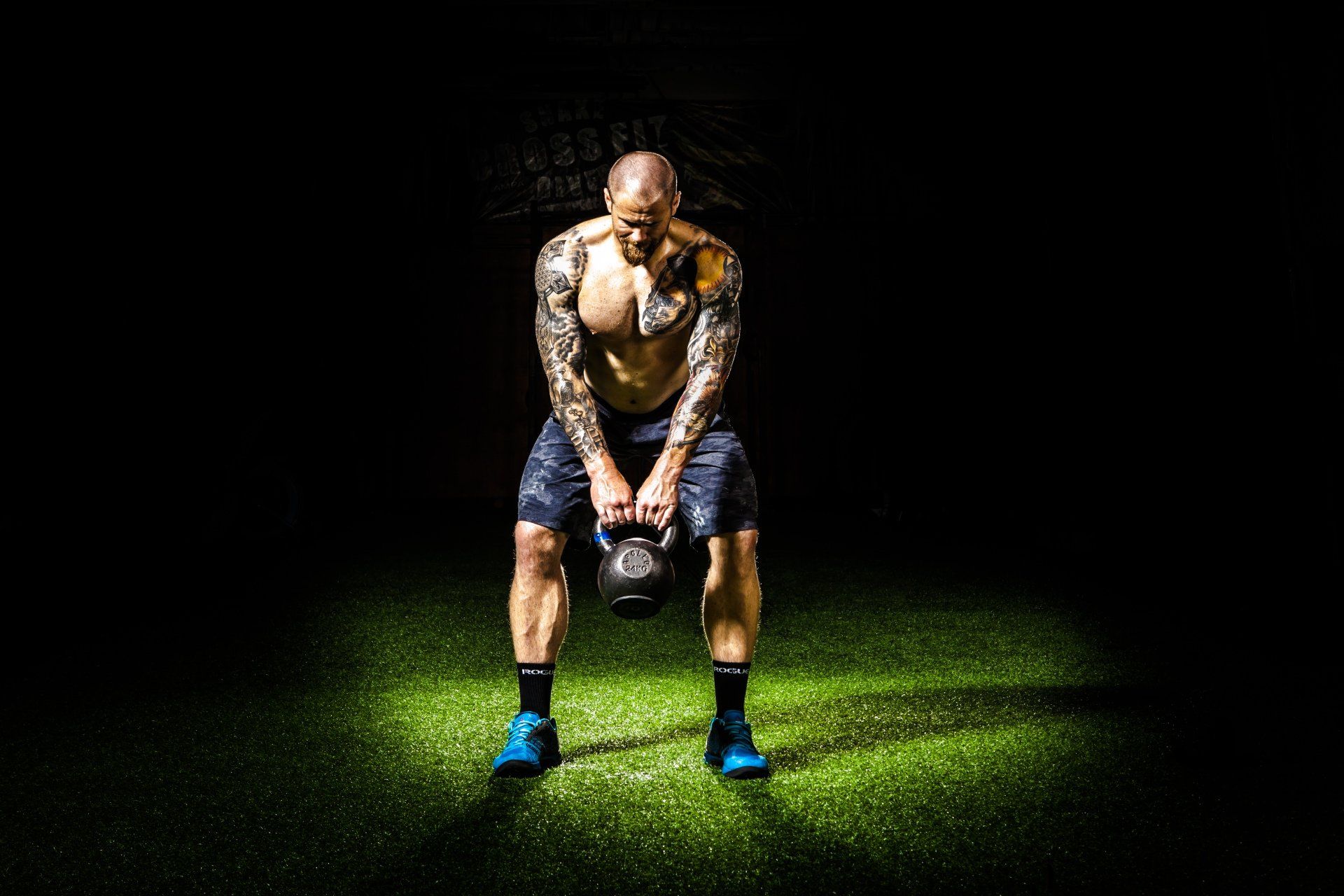 Man with tattoos doing a kettlebell workout on a green surface, black background.