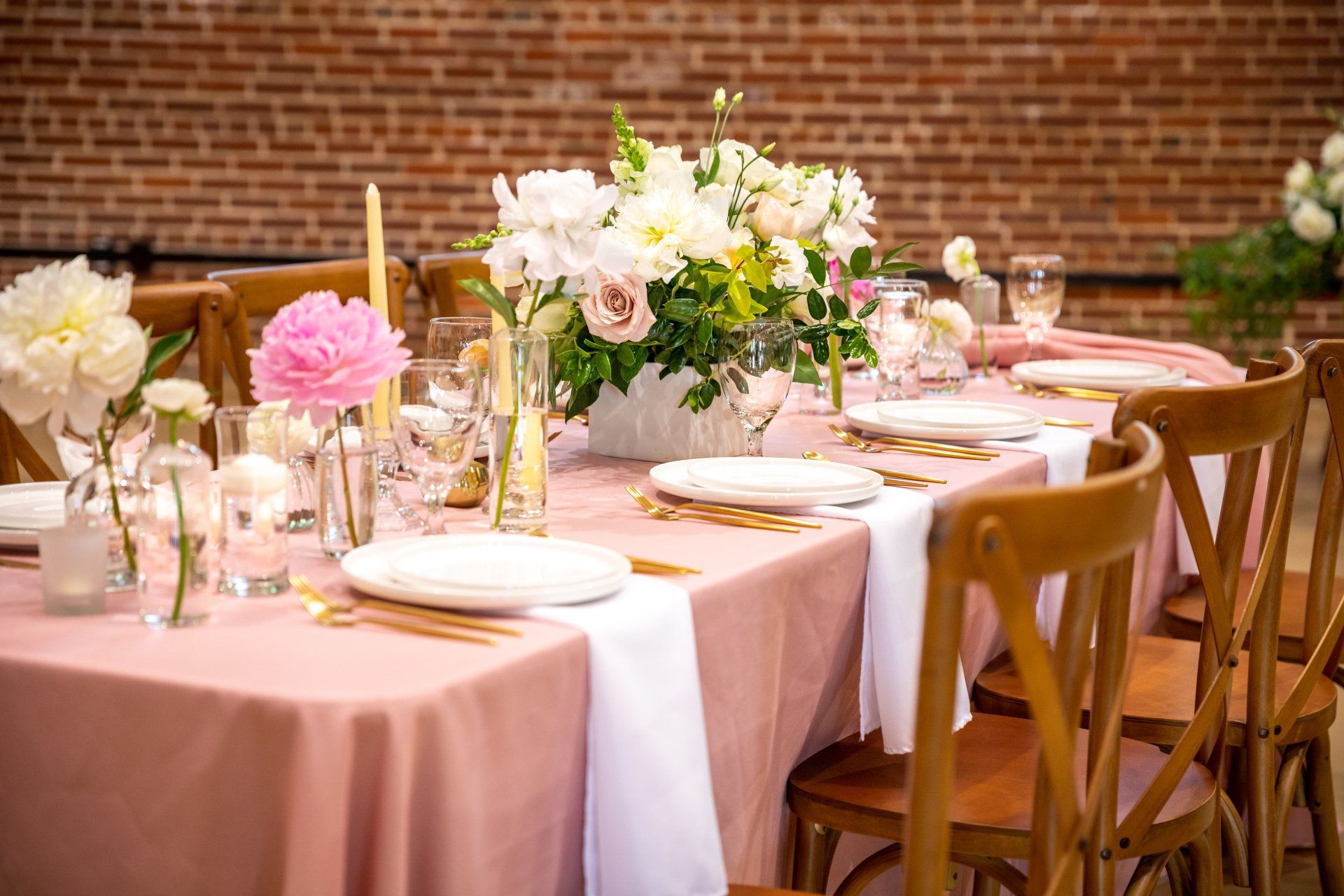 Formal table setting with pink tablecloth, flowers, and wooden chairs in a brick-walled room.
