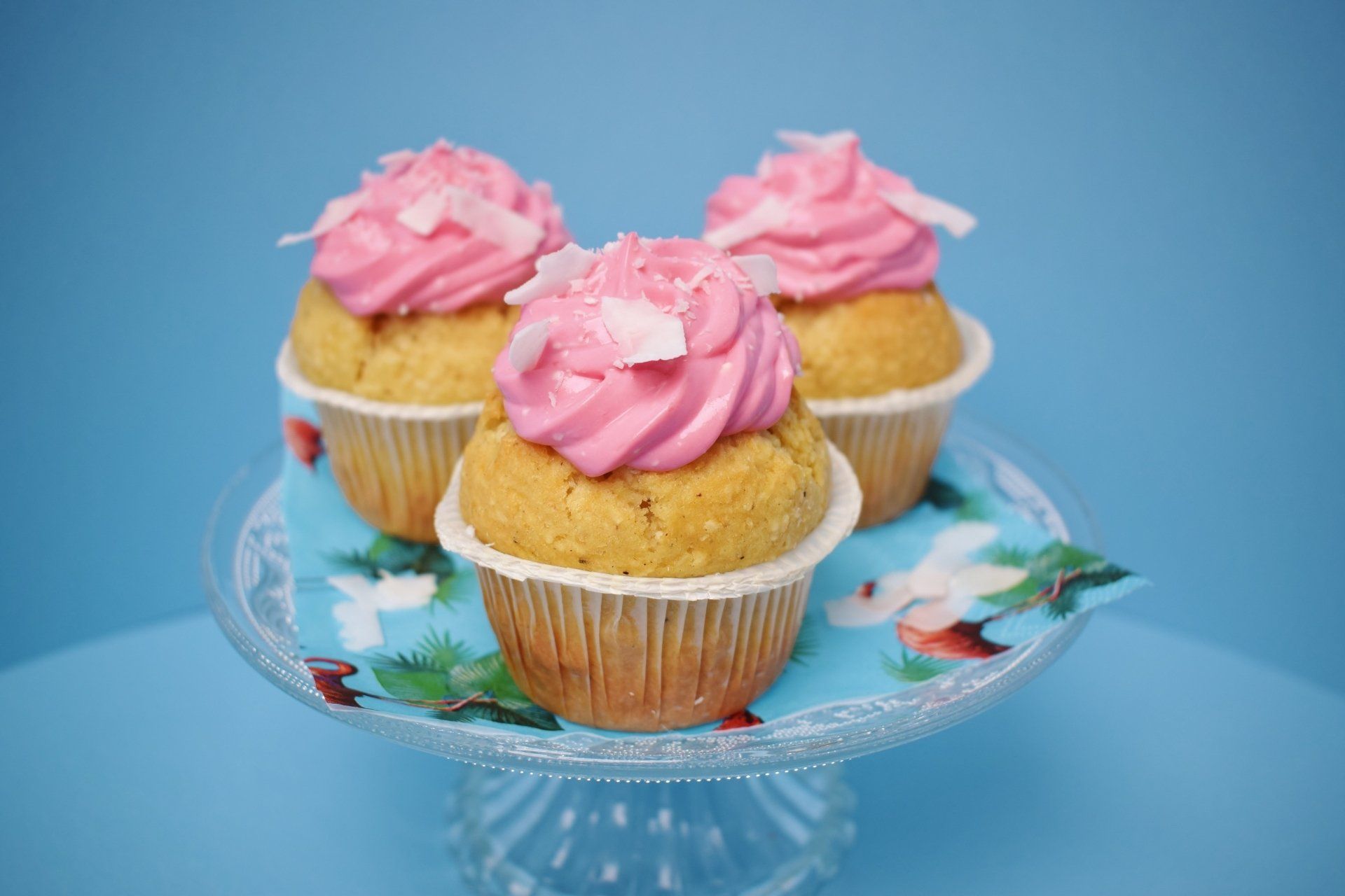 Three golden cupcakes topped with vibrant pink frosting and white coconut shavings on a glass stand against a blue background
