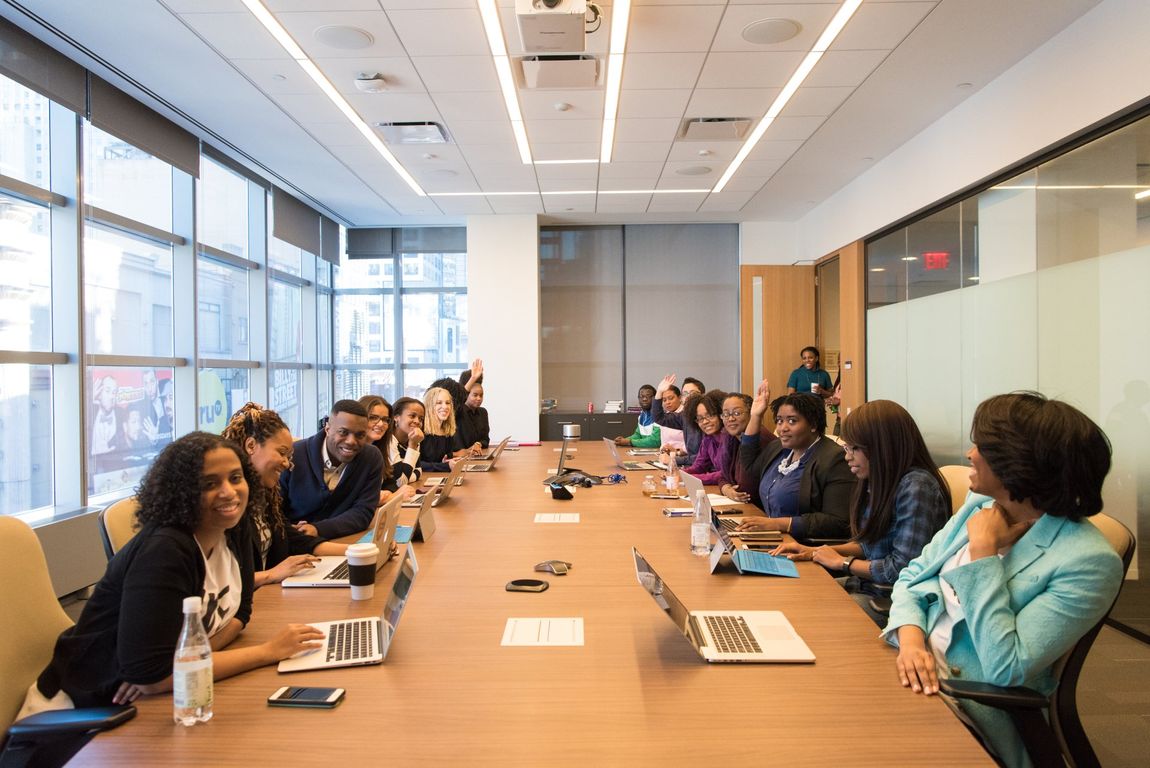 People in a meeting at a long table. Several are using laptops. Bright room with city view.
