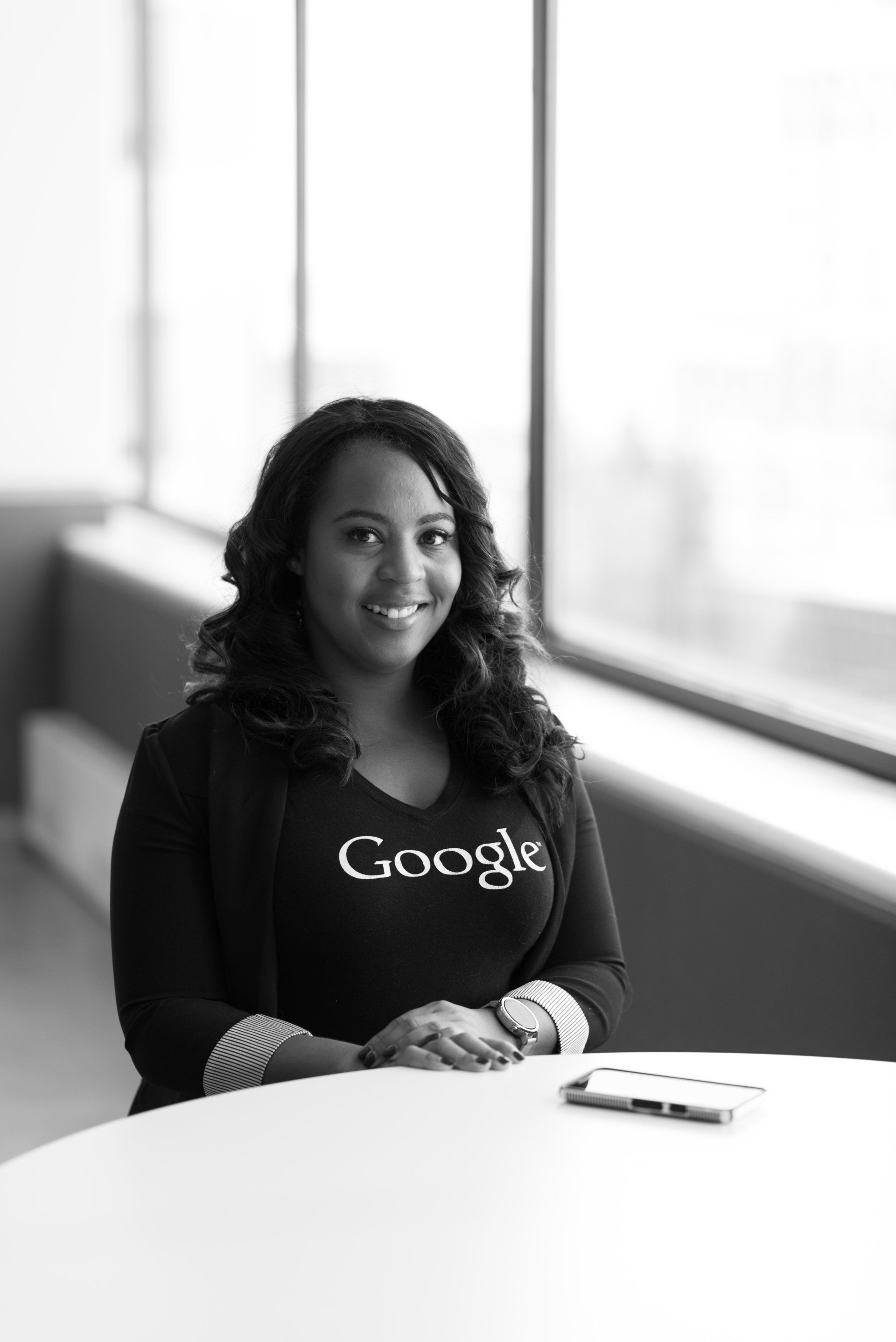 A woman wearing a google shirt is sitting at a table in front of a window.