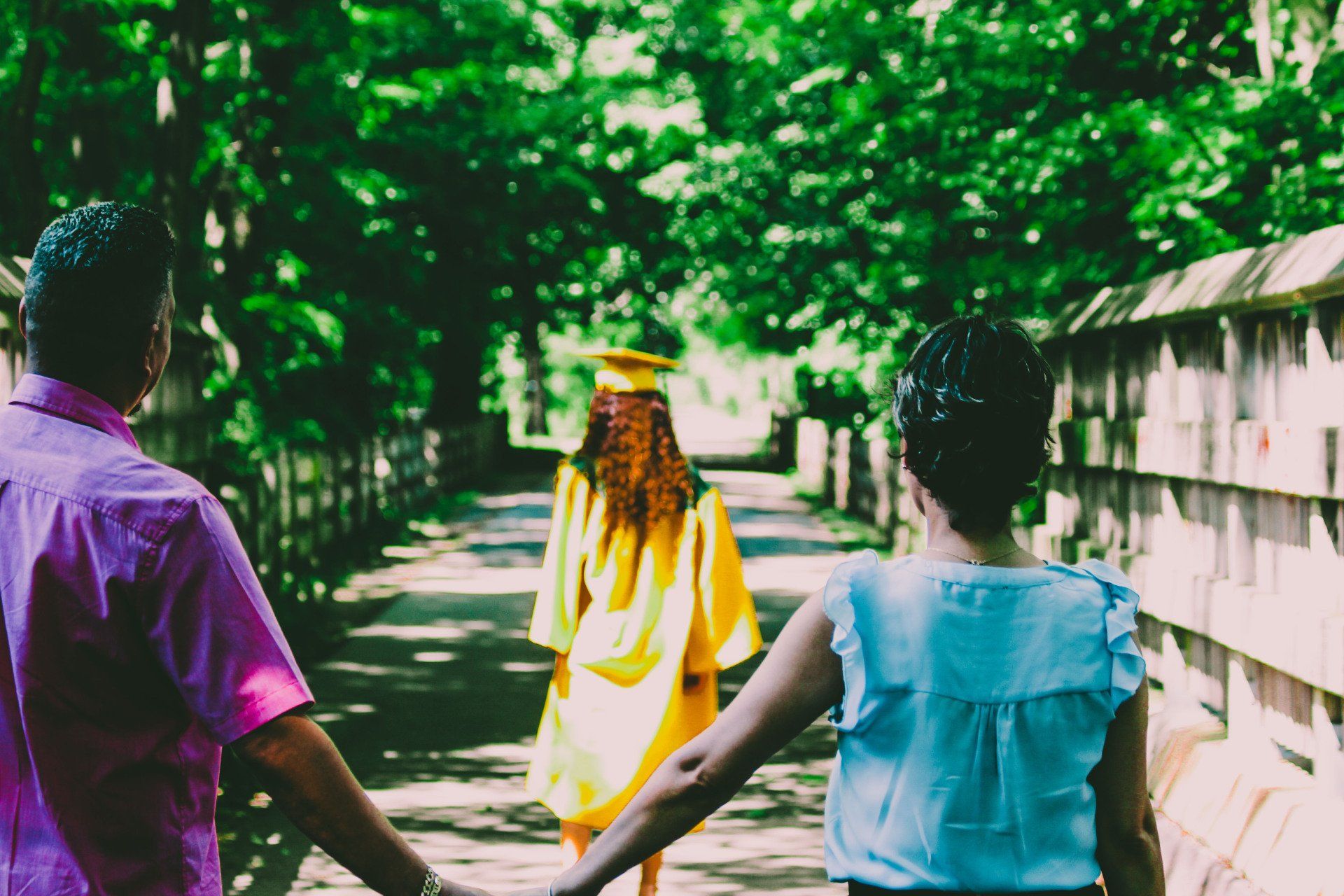 a woman in a graduation cap and gown is walking down a path with her parents holding hands .