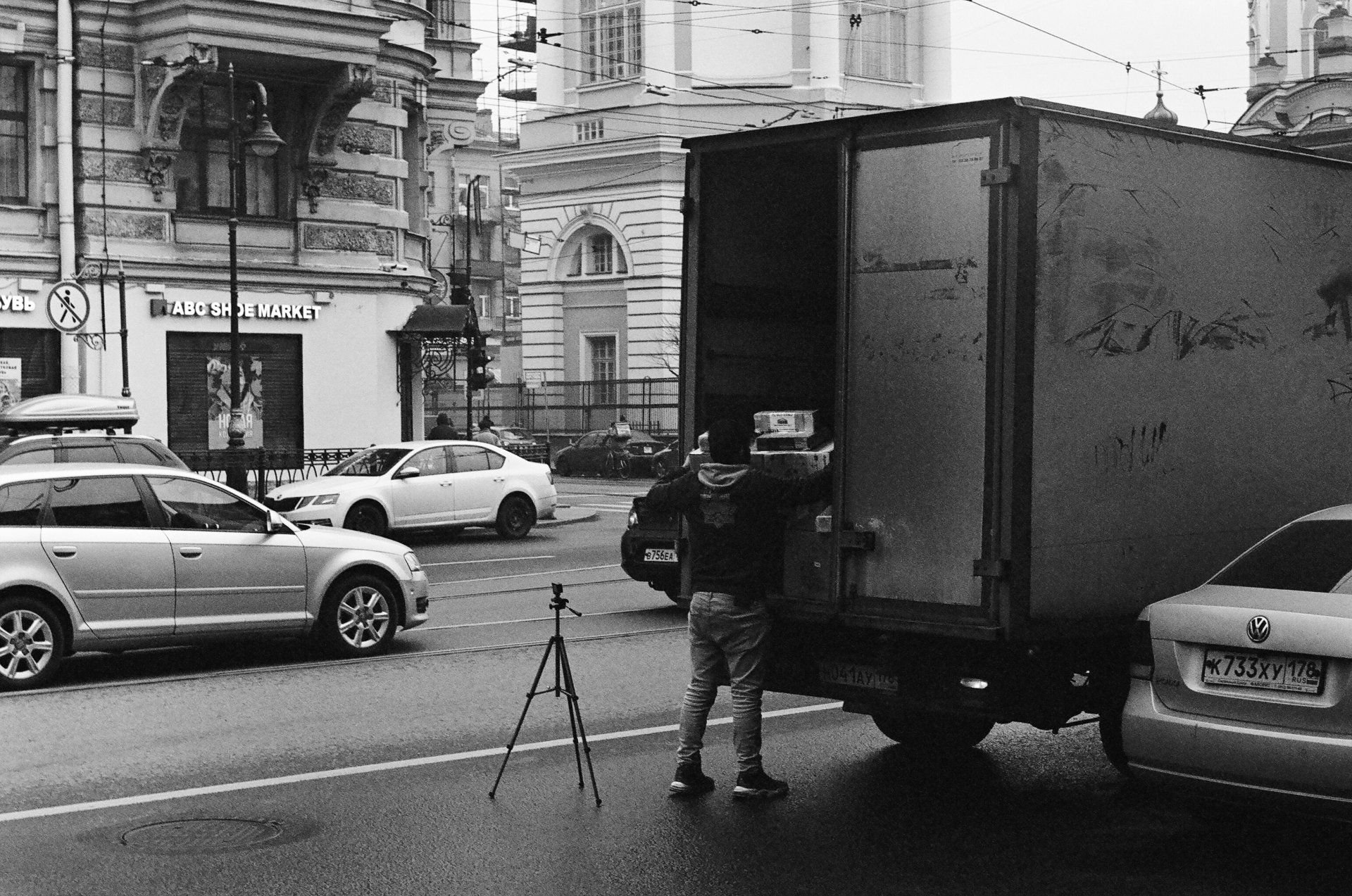 Person unloading a truck on a city street beside a parked car and tripod