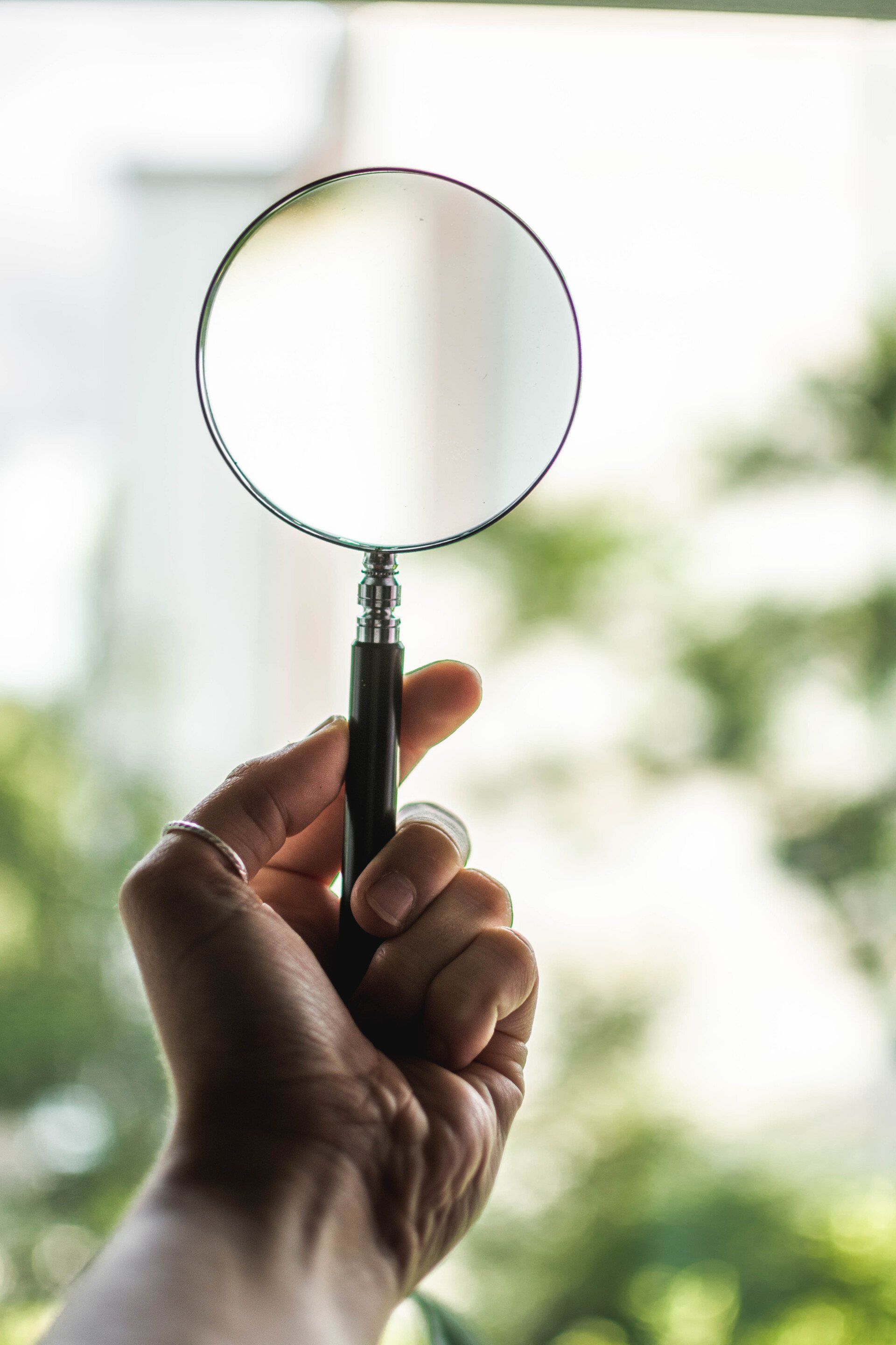Hand holding a magnifying glass against a bright window with blurred greenery outside