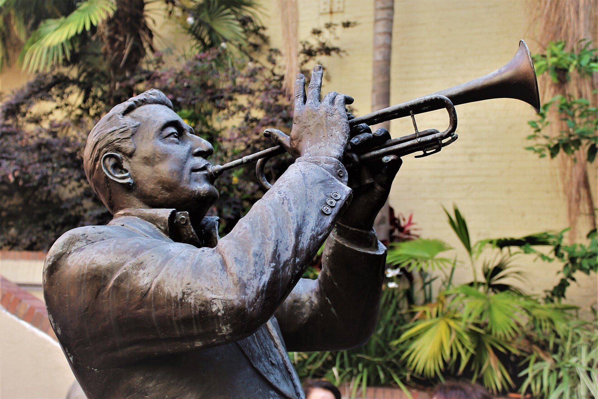Statue of a man playing the trumpet, bronze colored, outdoors, with green foliage in the background.