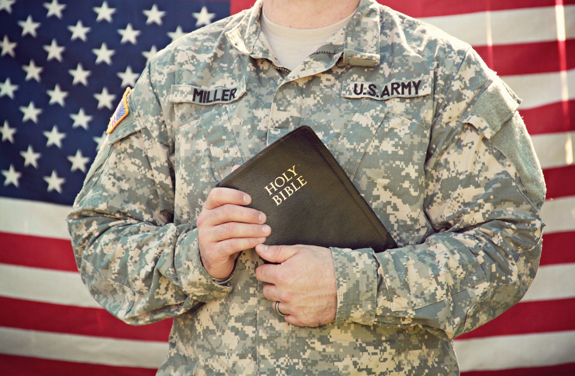 A soldier in camouflage uniform holding a Holy Bible in front of a United States flag.