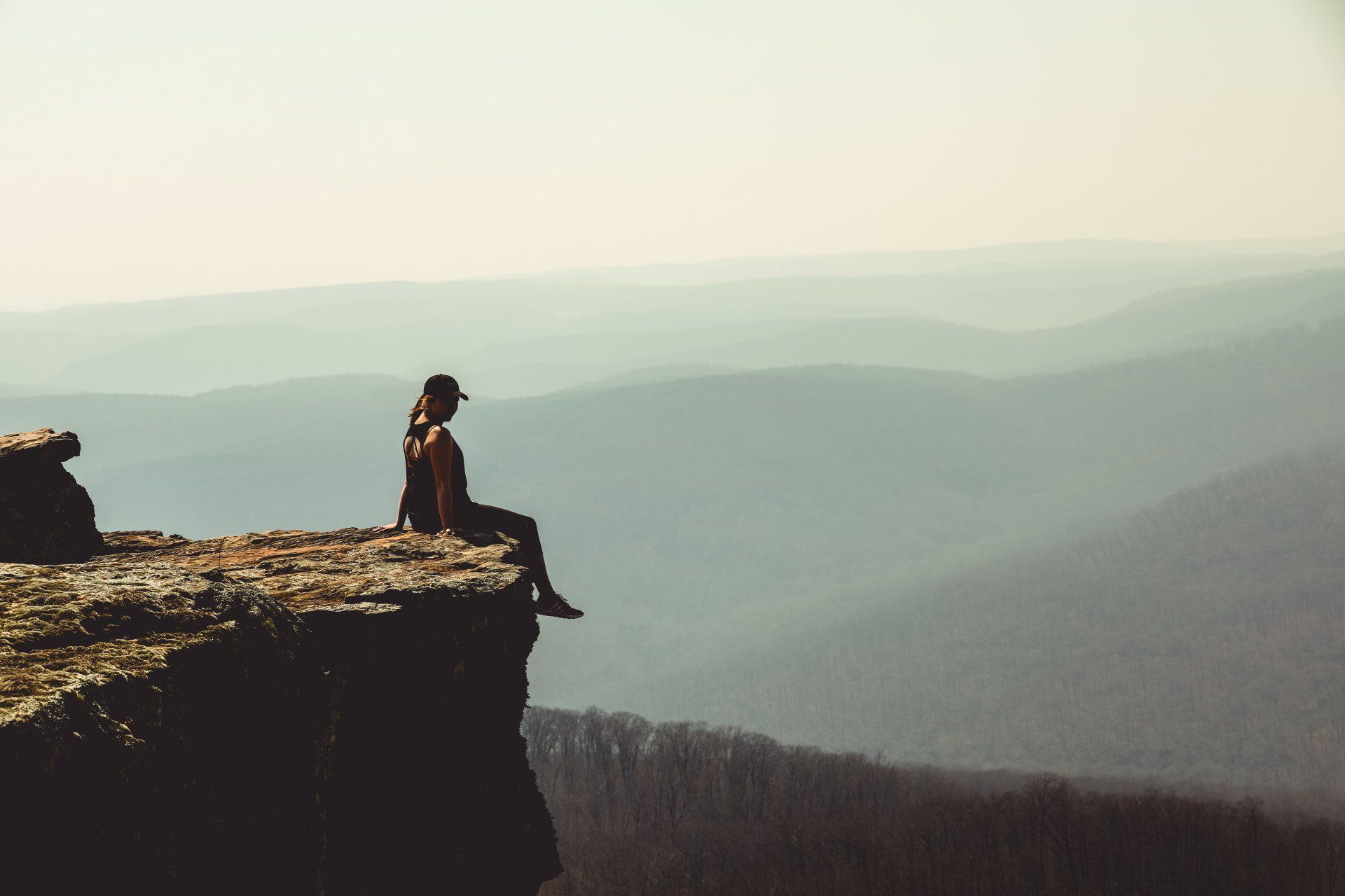 Person sitting on a cliff edge, looking out at a hazy mountain landscape.