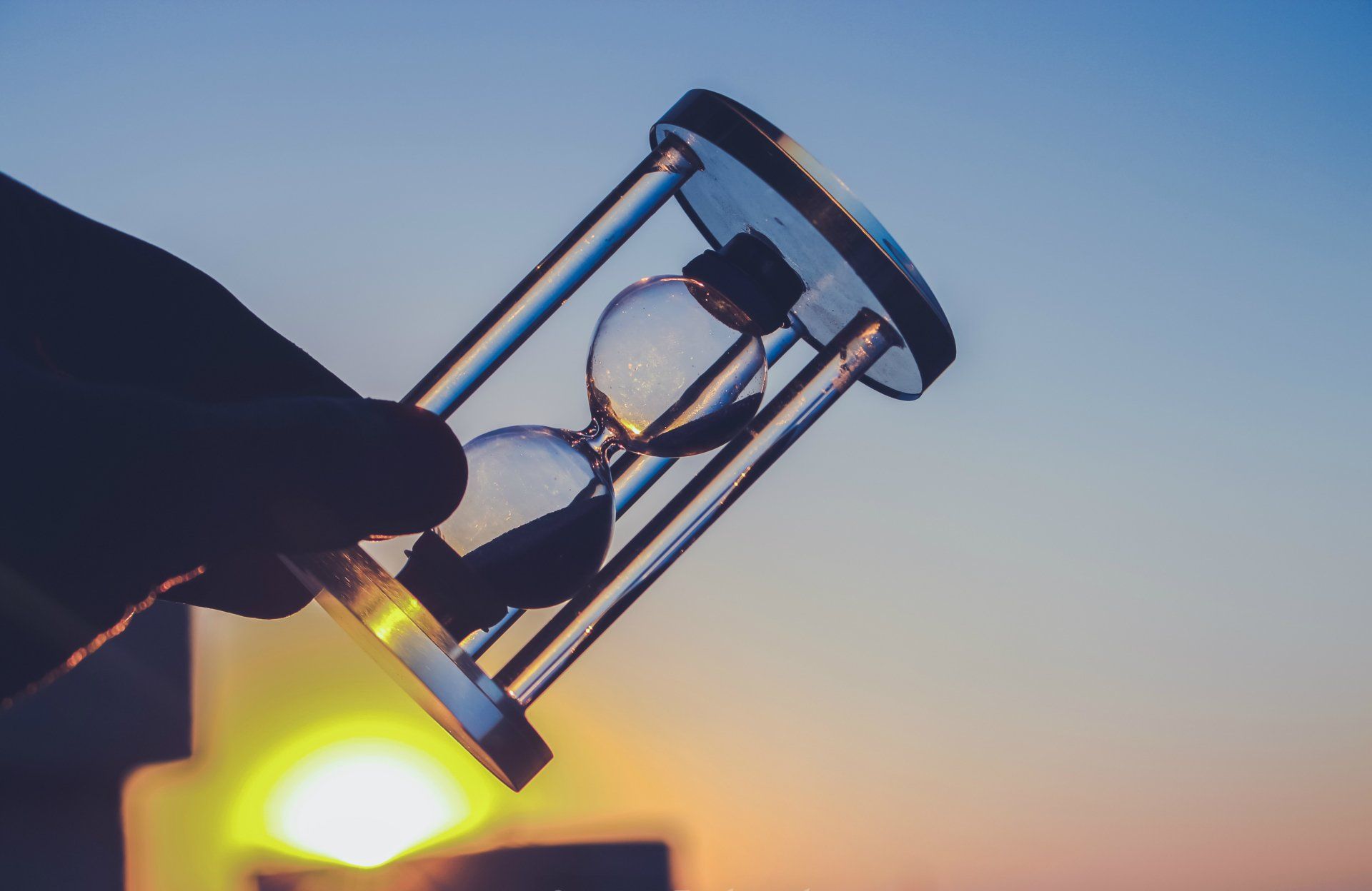 A hand is holding an hourglass in front of a sunset.