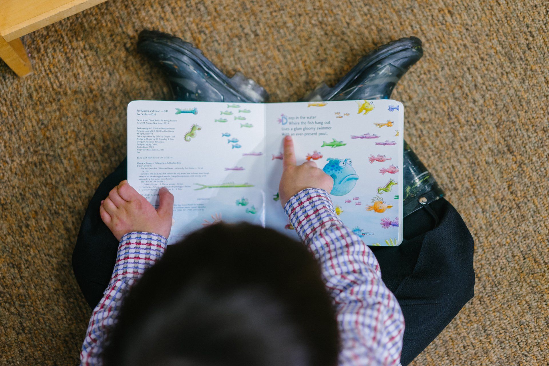 Child reading an open colorful book on a carpeted floor with legs crossed.