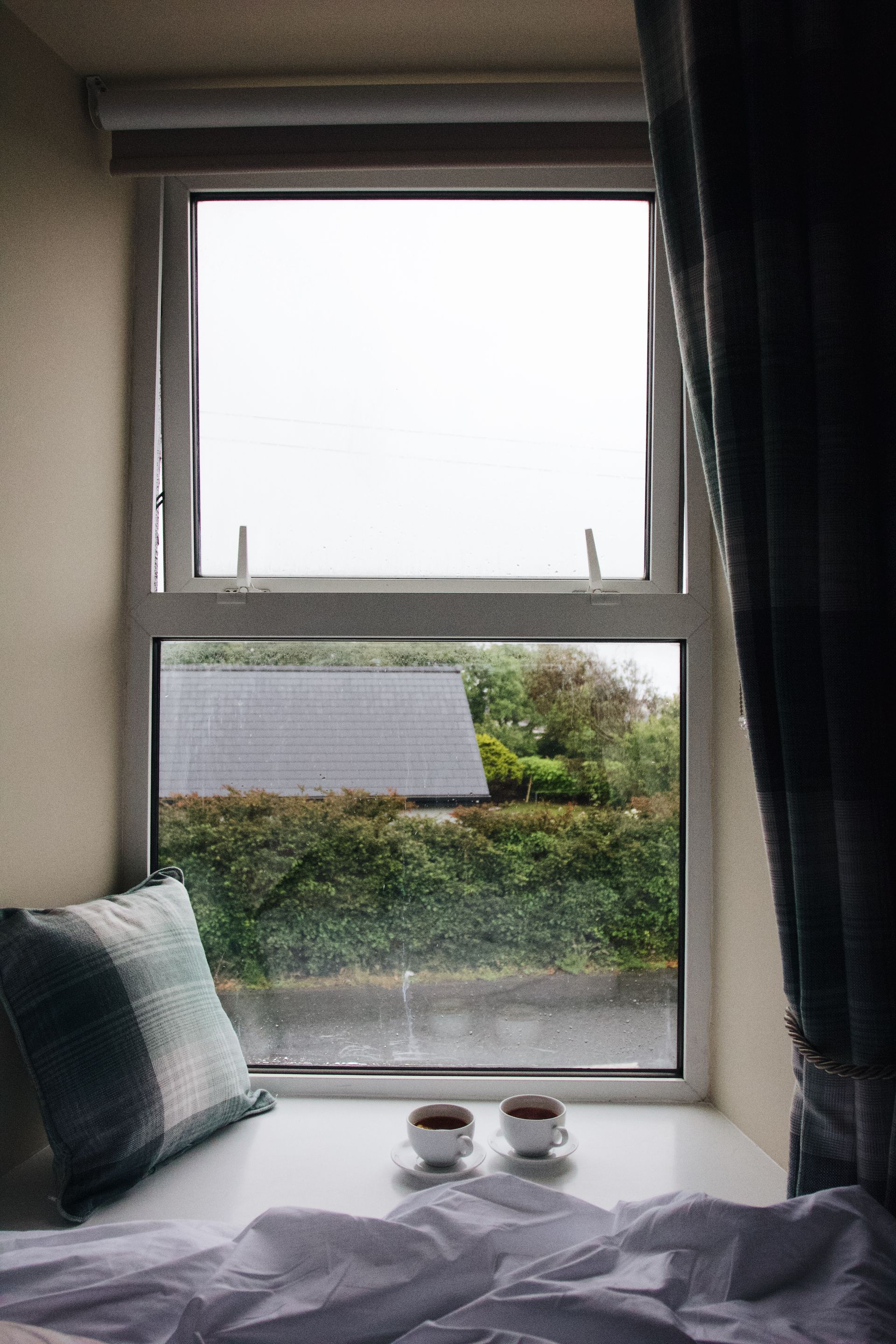 Cozy bedroom window view with greenery outside, bed in foreground, and curtains framing the scene.