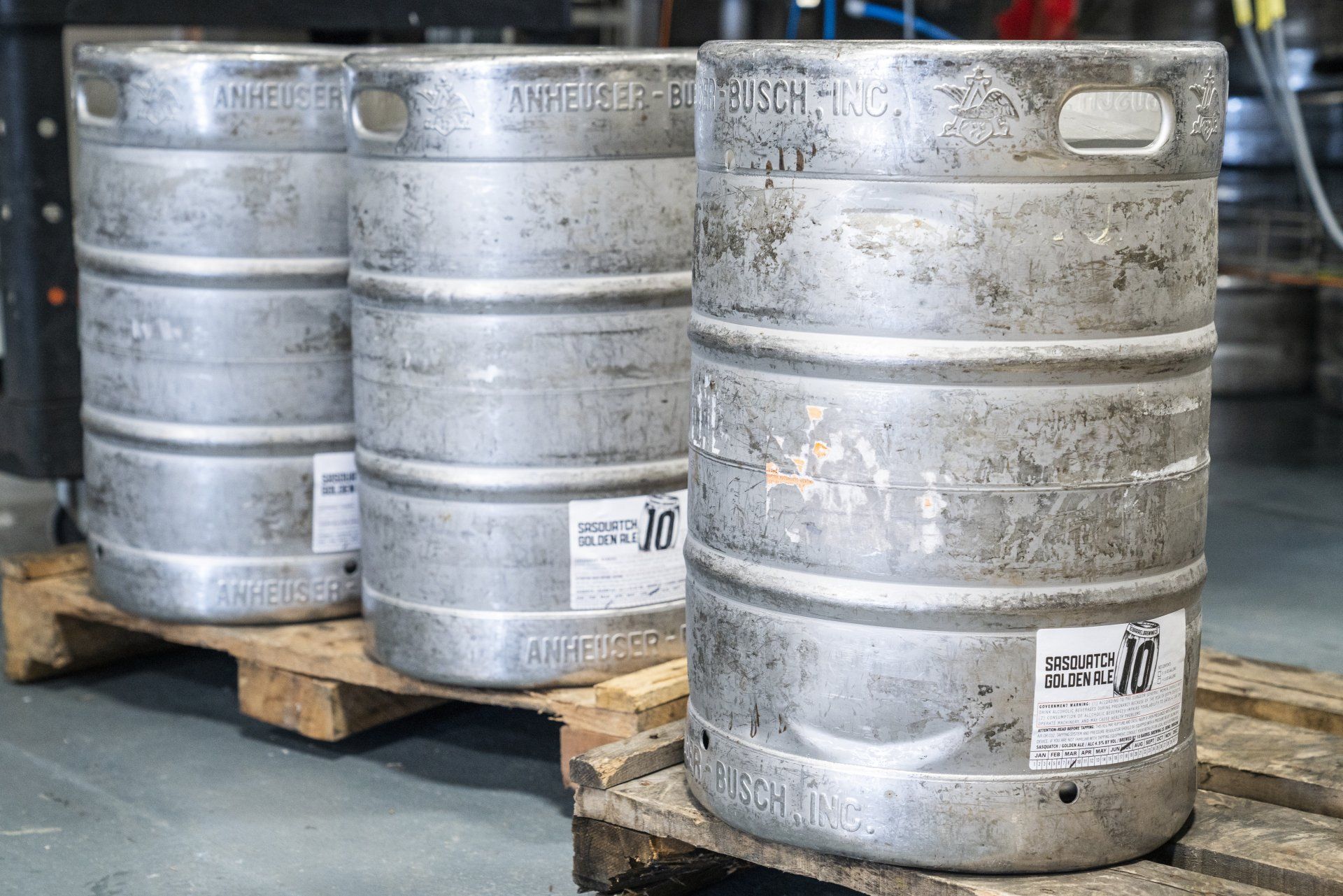 Three silver beer kegs on a wooden pallet in an industrial setting.