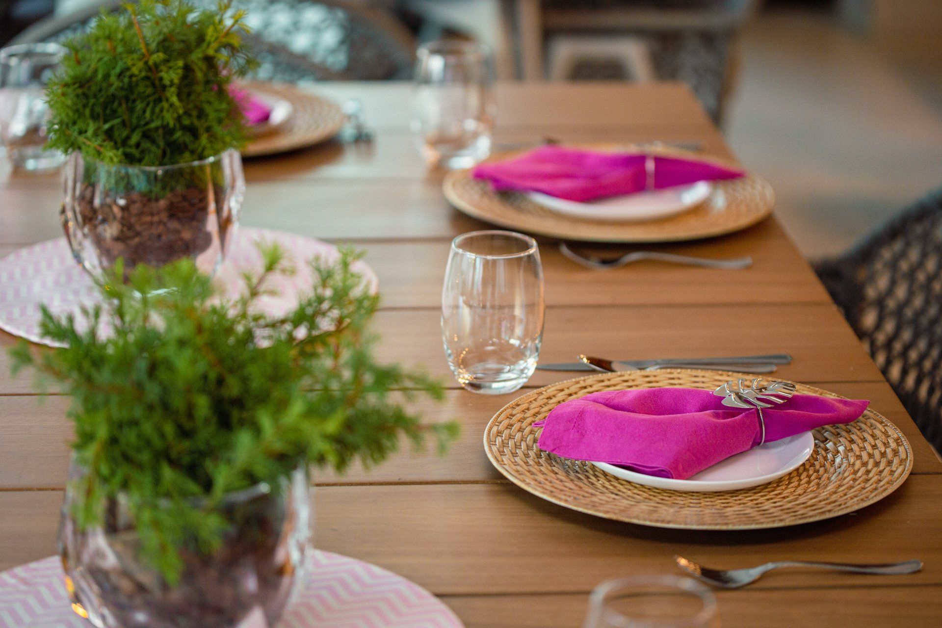 Table setting with pink napkins, white plates, gold chargers, clear glasses, and greenery.
