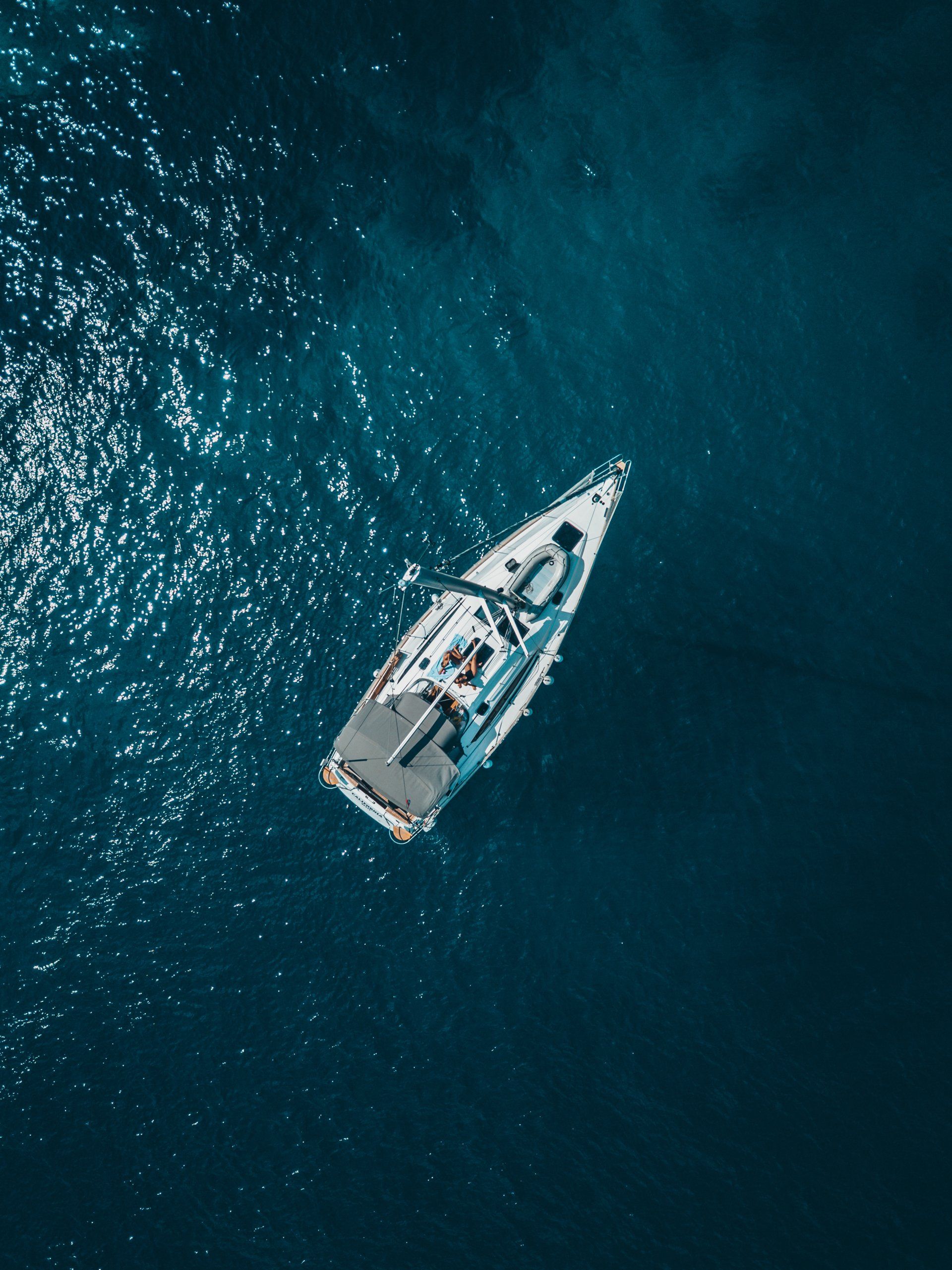 An overhead aerial view of a white sailboat drifting on deep blue, sparkling ocean water.