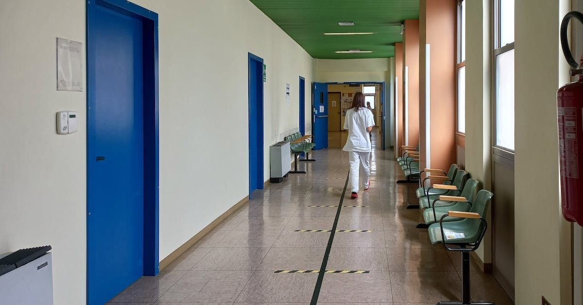 Hospital corridor with blue doors, green ceiling, row of chairs, and a person walking away.