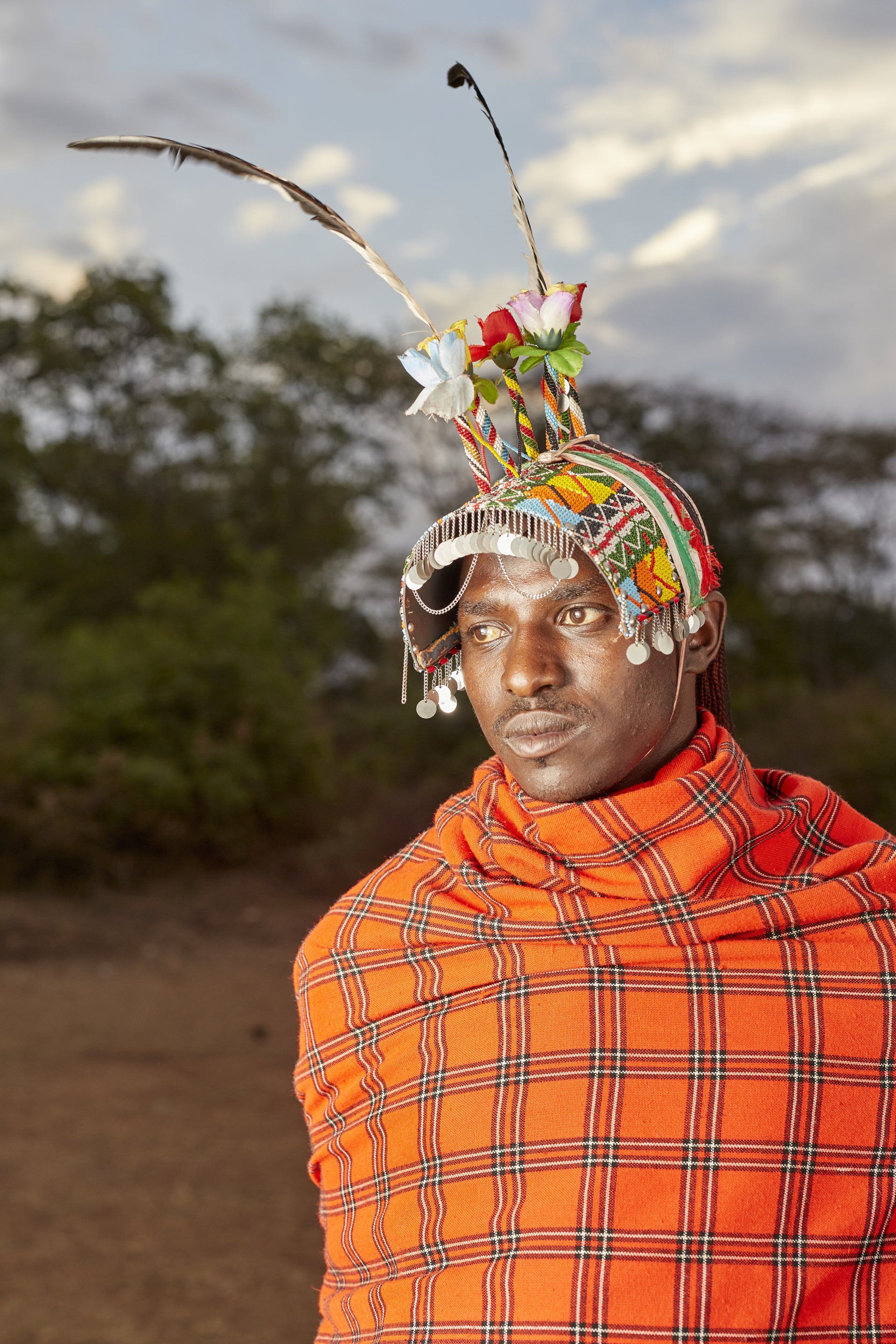 Man wearing traditional beaded headdress and red plaid garment, outdoors.